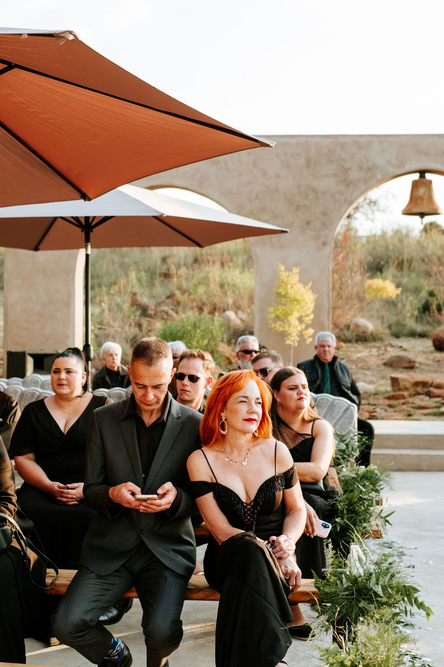 Wedding guests in black attire seated outdoors under terracotta umbrellas during ceremony.
