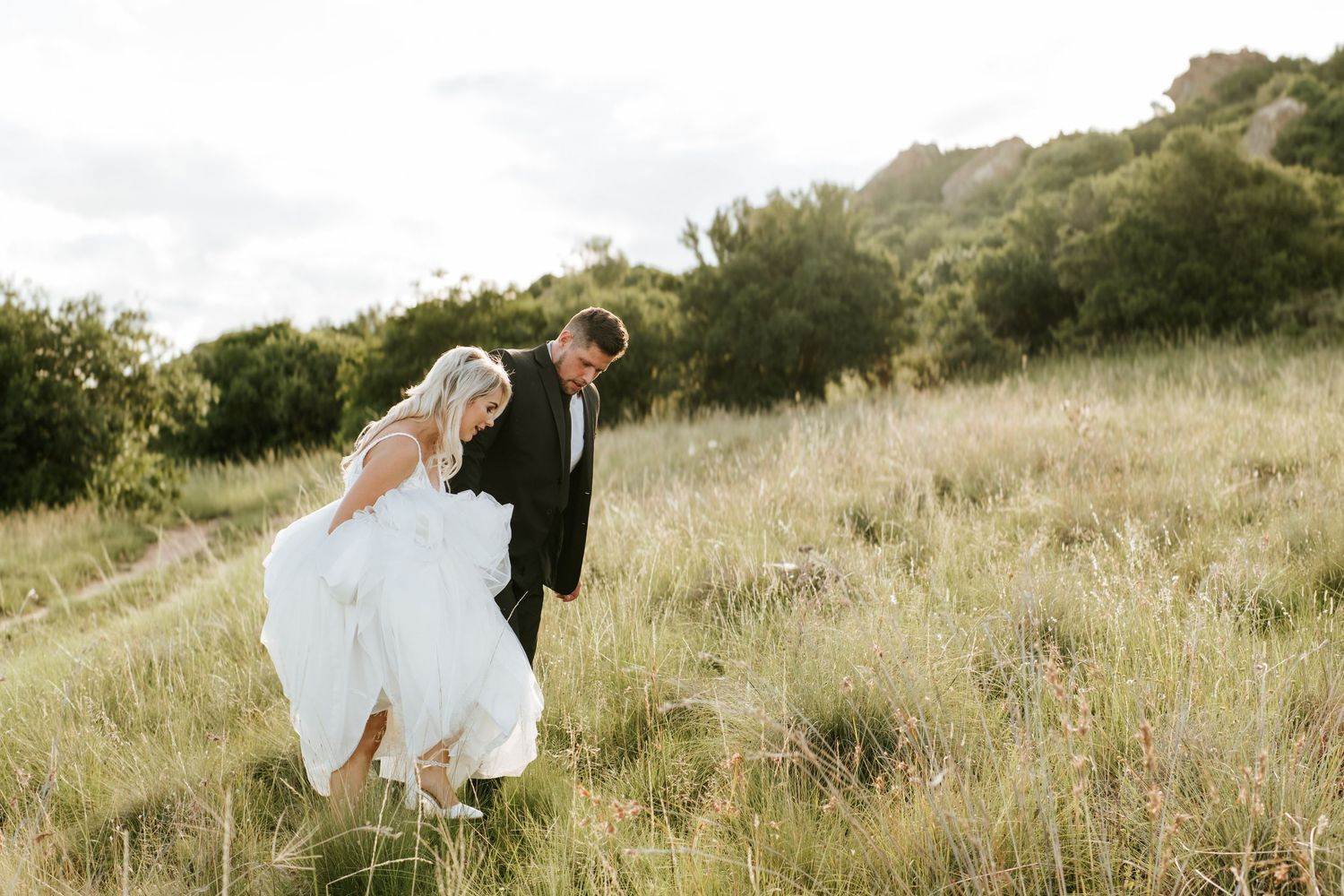 Smiling bride lifts her white gown while walking through tall golden grass with her groom.