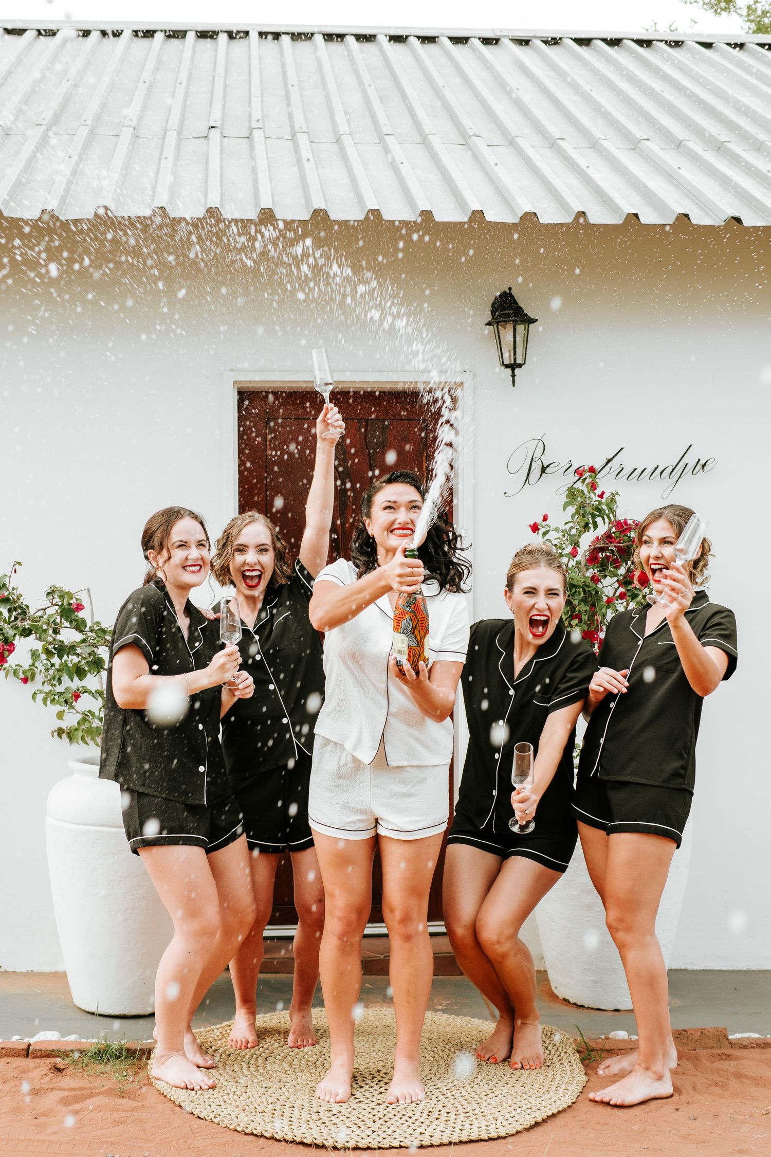 Bride and bridesmaids in black pajamas celebrate with champagne spray outside a white farmhouse building.