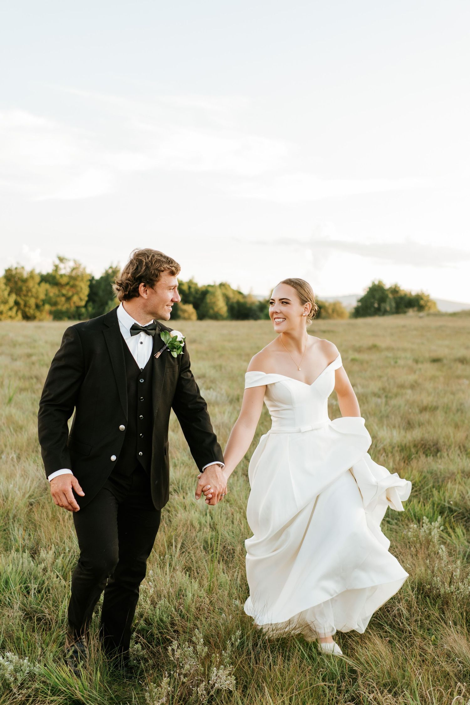 Bride and groom holding hands walking through a green meadow, bride in off-shoulder gown, groom in black tuxedo.