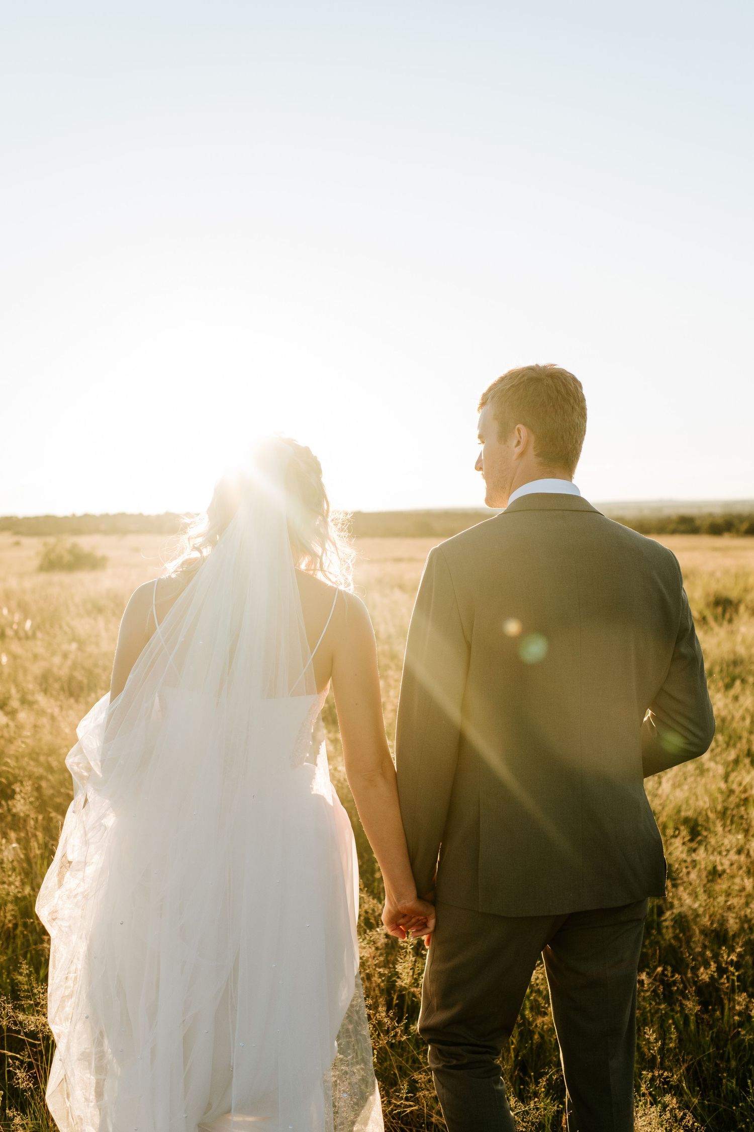 Bride and groom holding hands walking through golden sunlit field, bride wearing flowing white gown with veil.