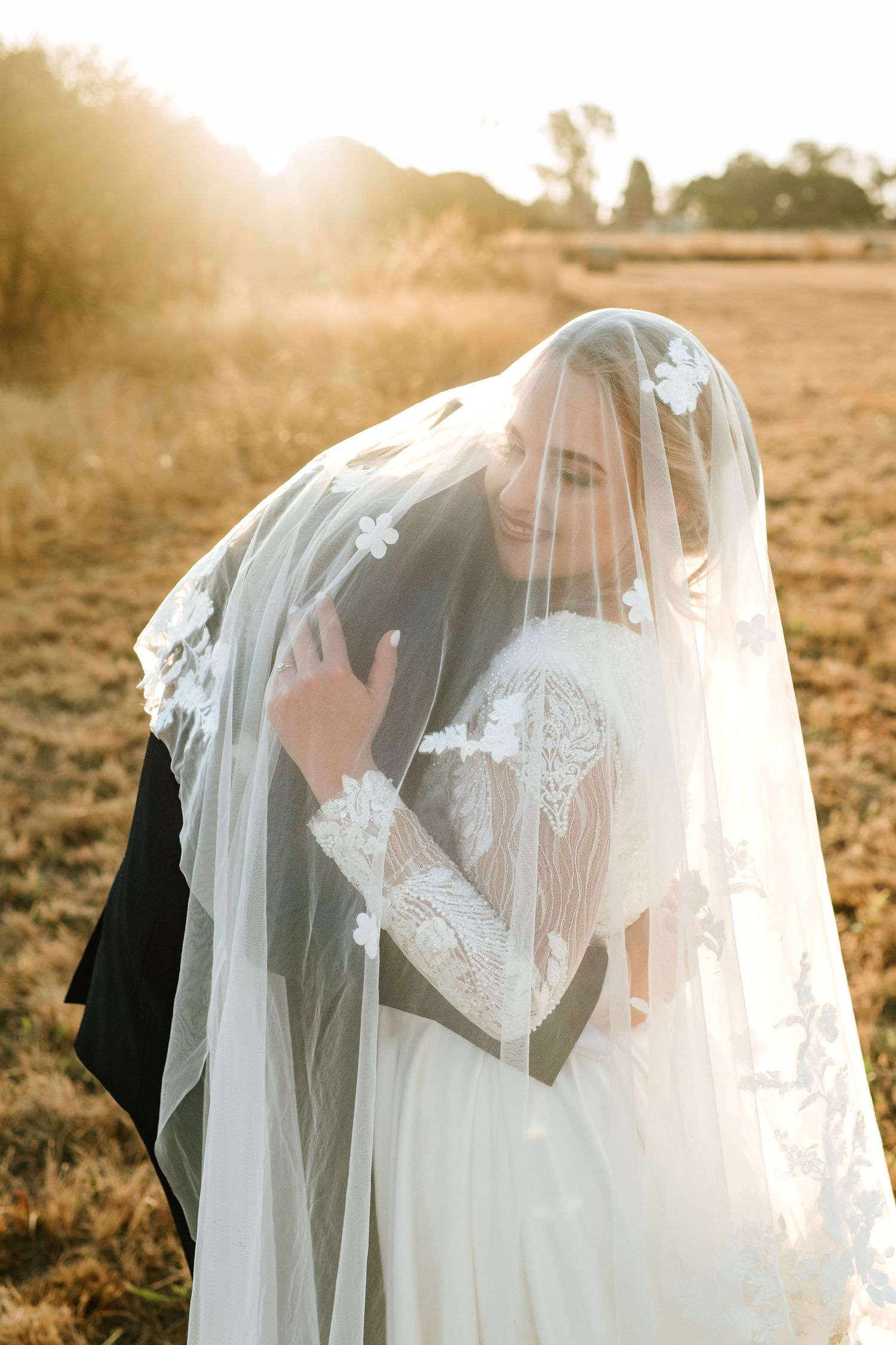 Bride and groom share an intimate moment under a floral veil in a golden sunset field.