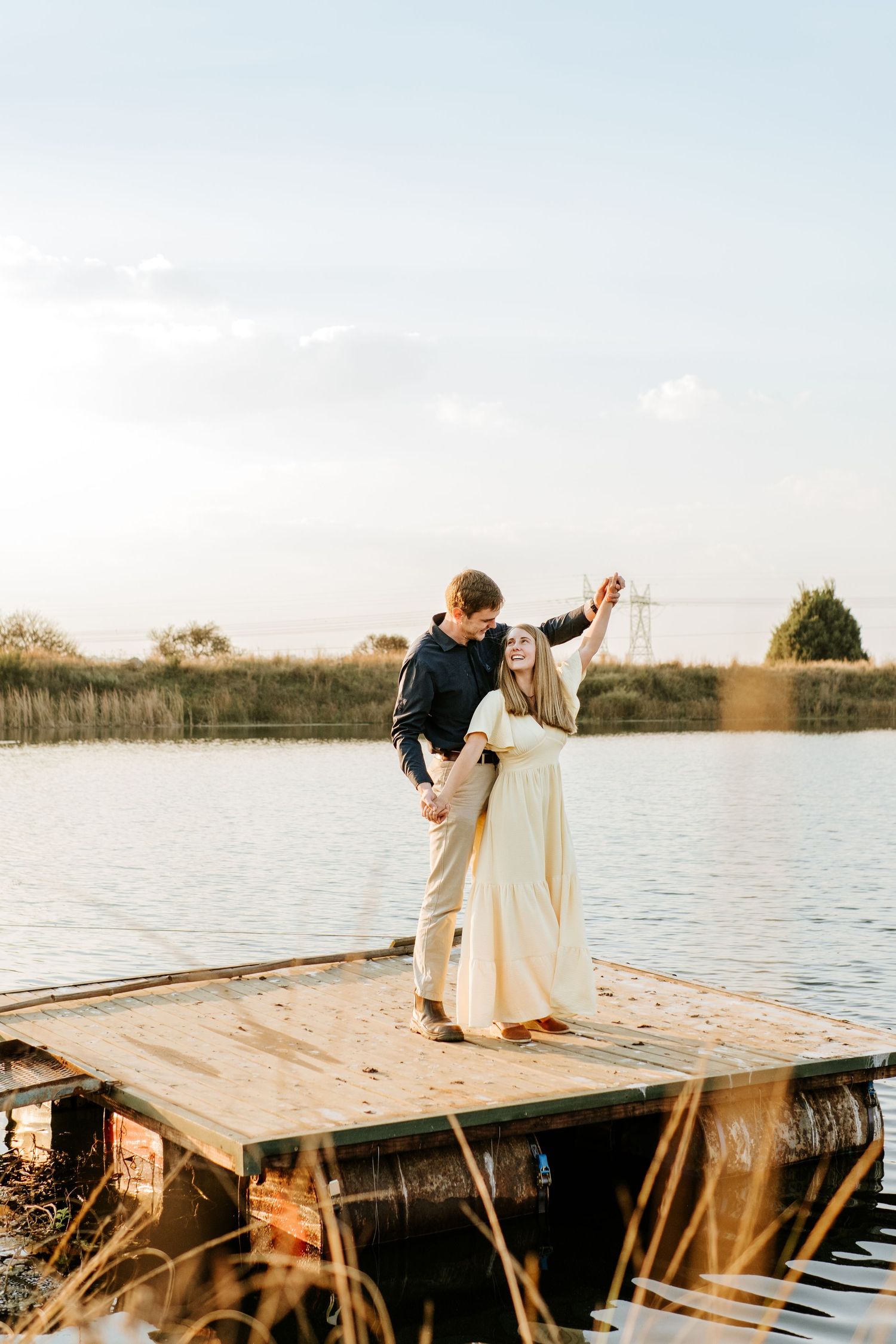 Joyful couple dancing together on a wooden dock over a calm lake during a beautiful golden sunset.