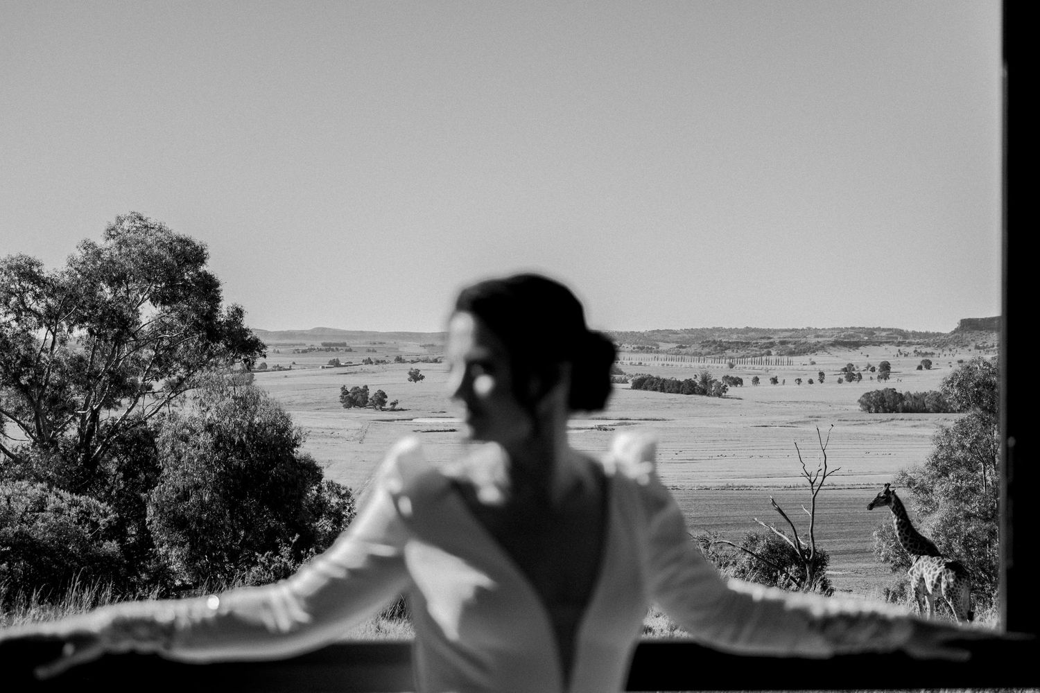Black and white photo of a bride in a wedding dress overlooking a vast rural landscape from a balcony.
