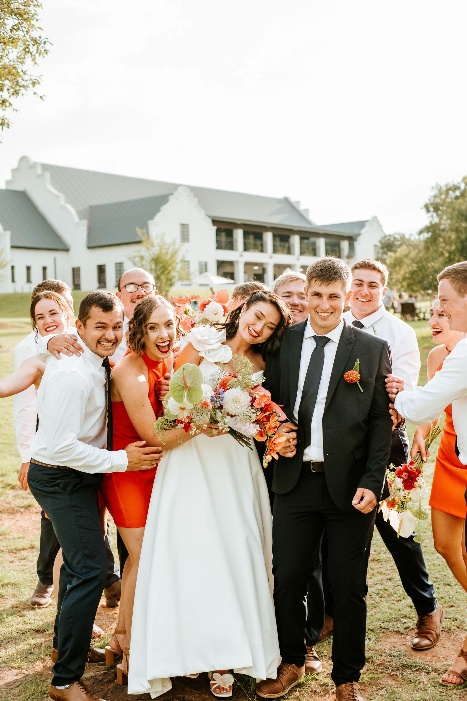 Joyful wedding party group photo with bride, groom, and guests in orange attire before white venue.