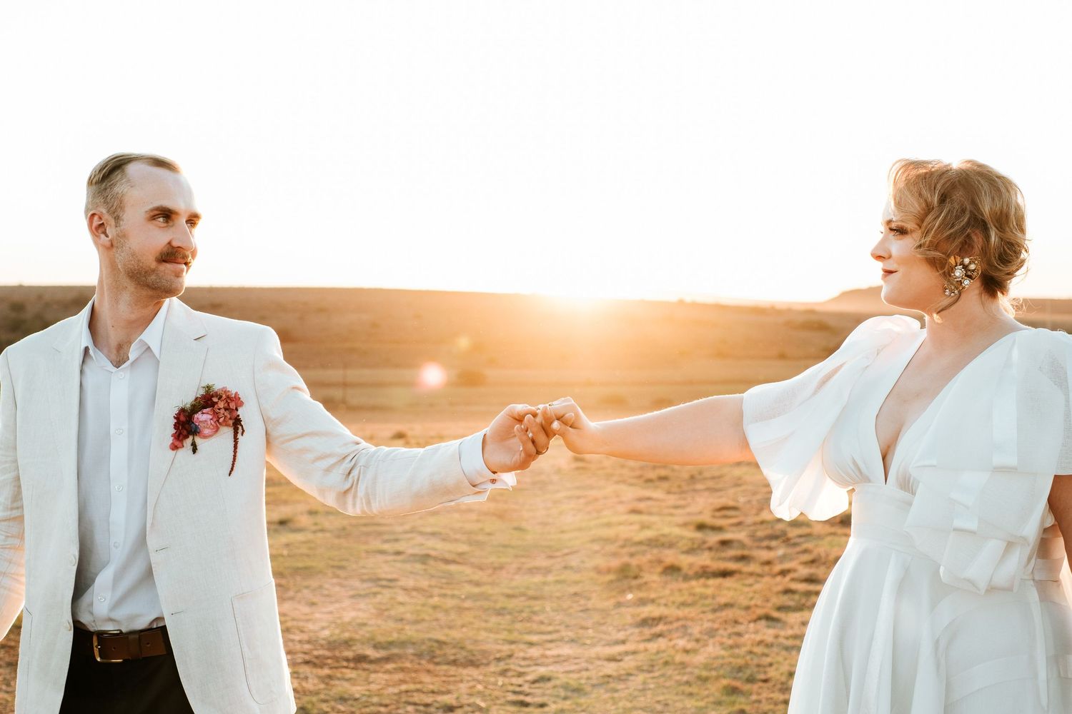 Bride and groom hold hands at arm's length in golden sunlit open field, smiling warmly photographed by Laughing Lamp Photo & Film