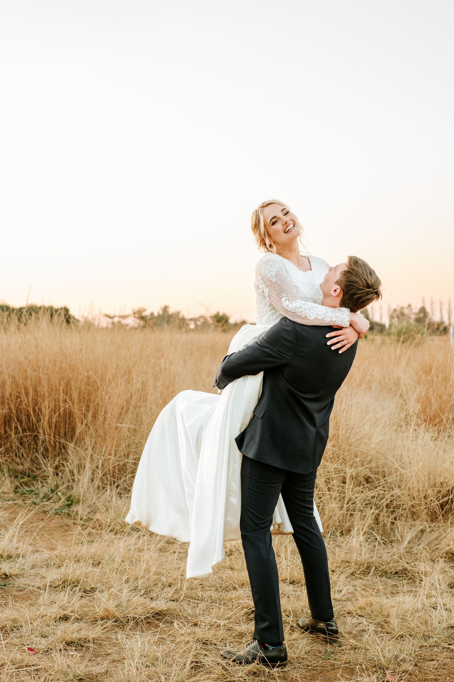 Joyful groom lifting smiling bride in a white dress in a golden wheat field at sunset.