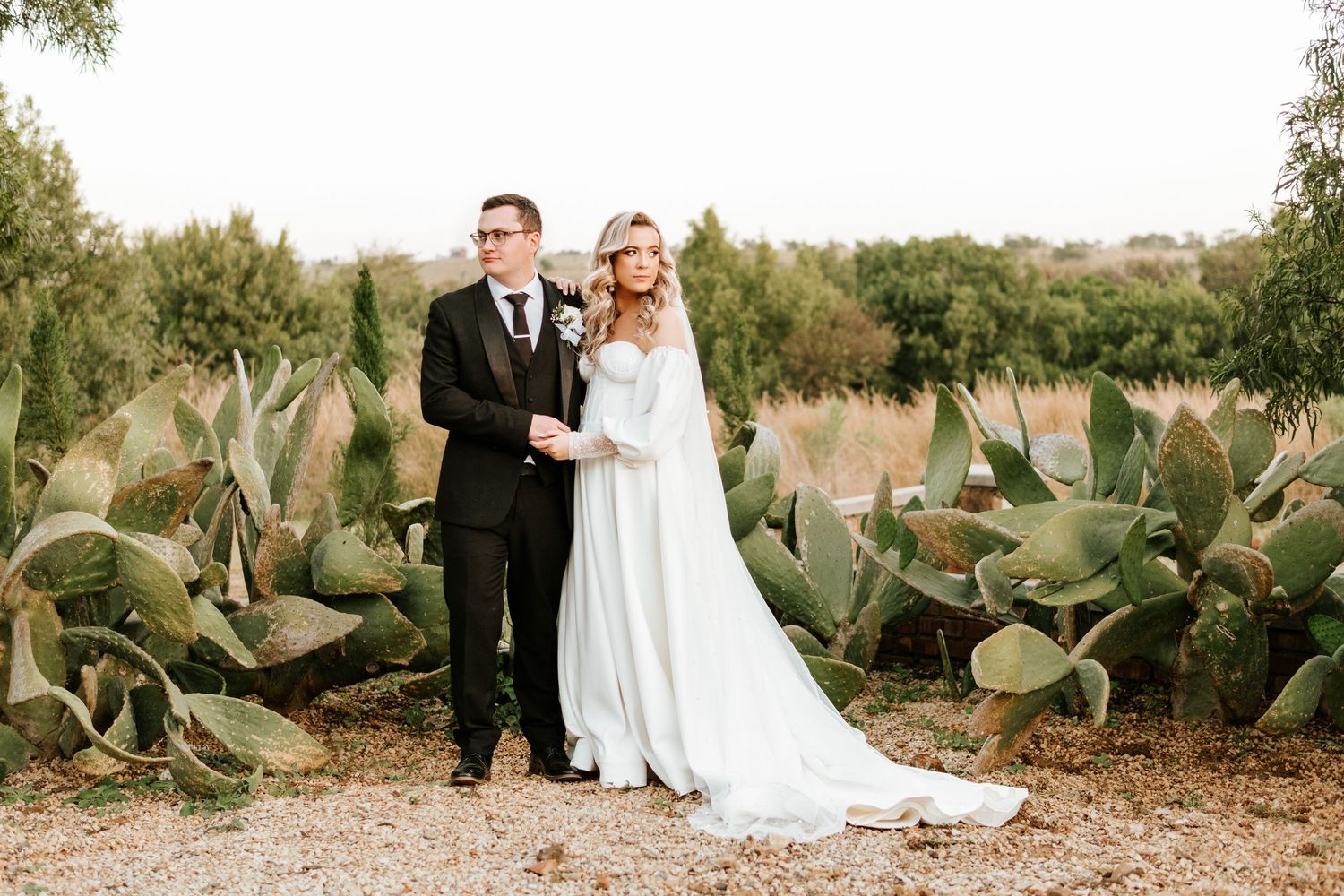 Bride and groom standing together among tall cacti in a scenic outdoor wedding portrait.