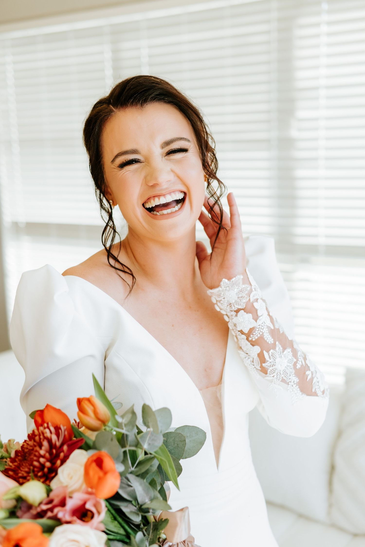 Radiant bride laughing joyfully while holding a colorful orange and red bouquet in bright indoor setting.