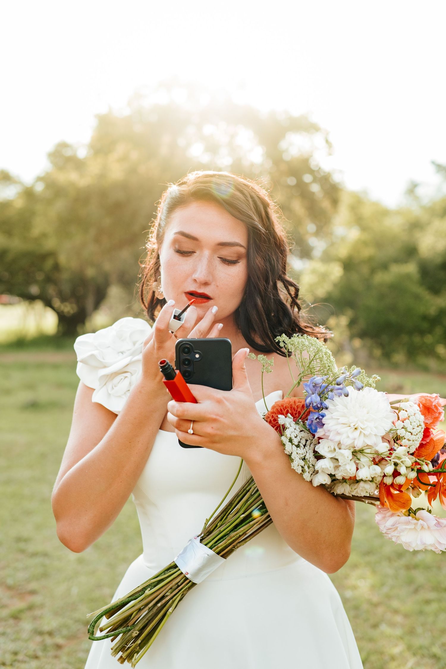 Bride in white dress touches up lipstick using phone camera while holding colorful bouquet.