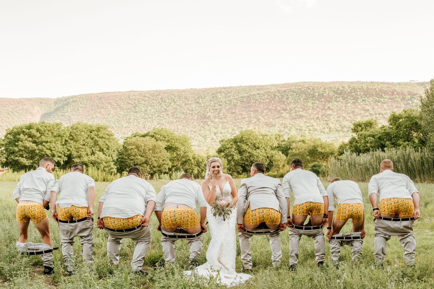 Bride stands in field as groomsmen in matching outfits bow down in a humorous pose.