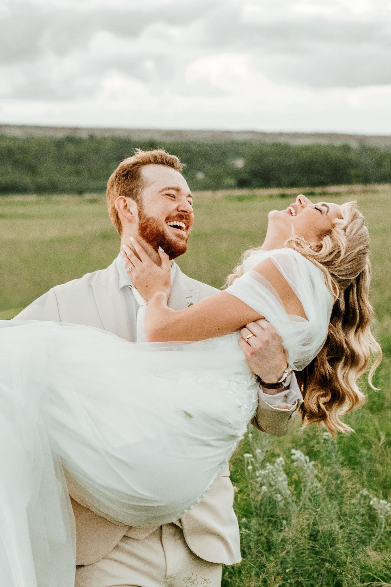 Laughing groom carries joyful bride in white gown through a lush green countryside field.