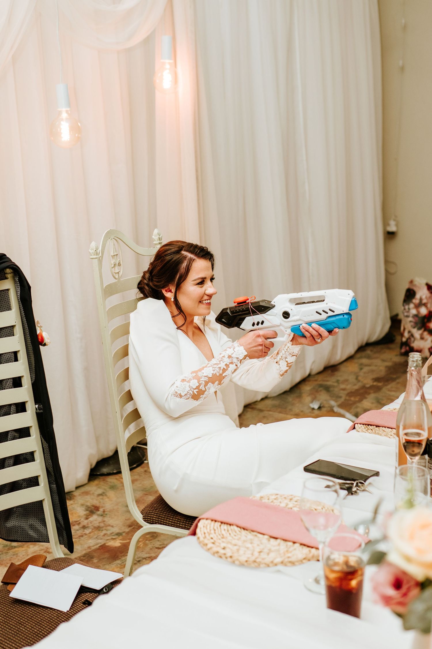 Smiling bride in white suit playfully holding a toy gun at wedding reception table.