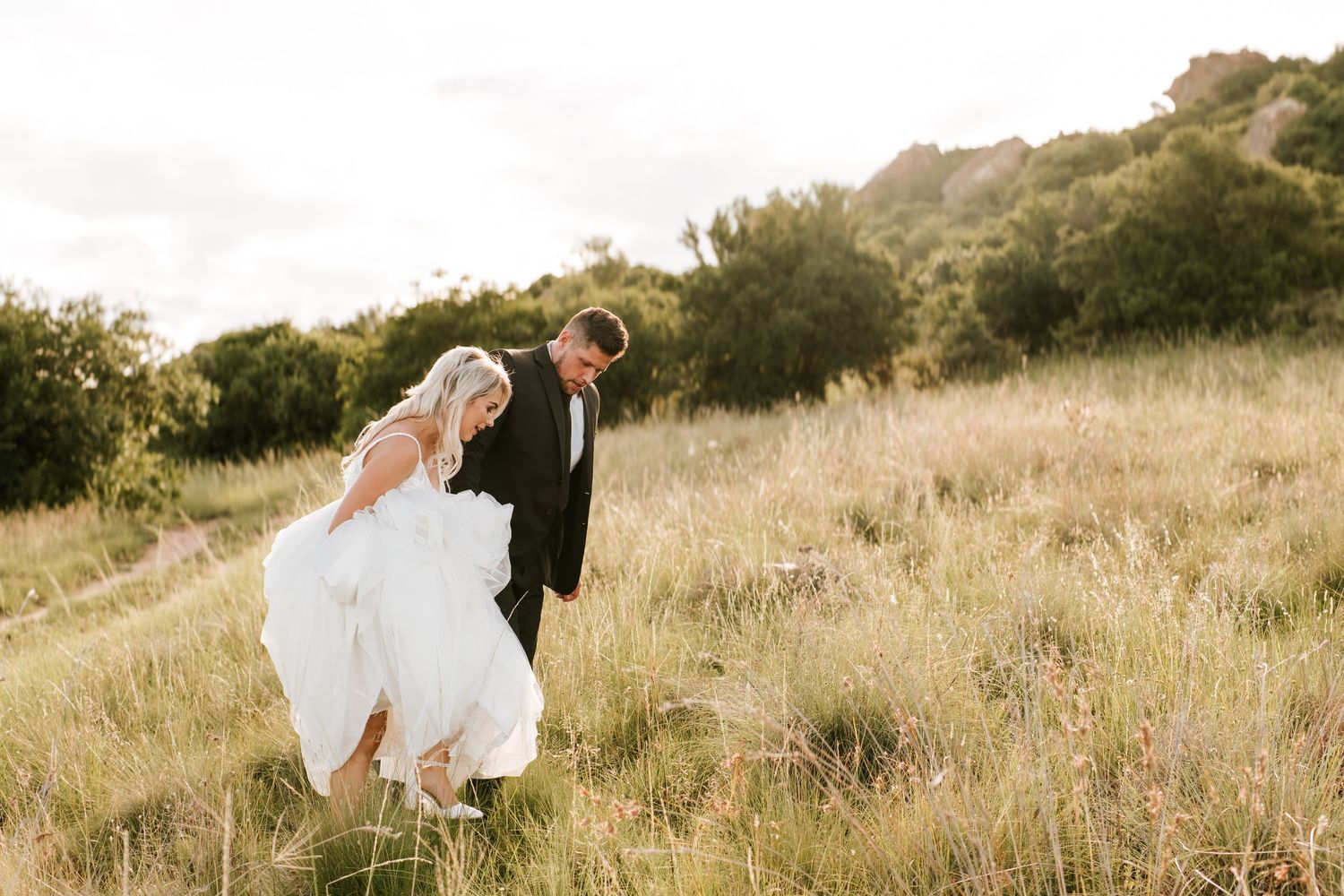 Bride in flowing white gown and groom in black tux walk through golden meadow at sunset.