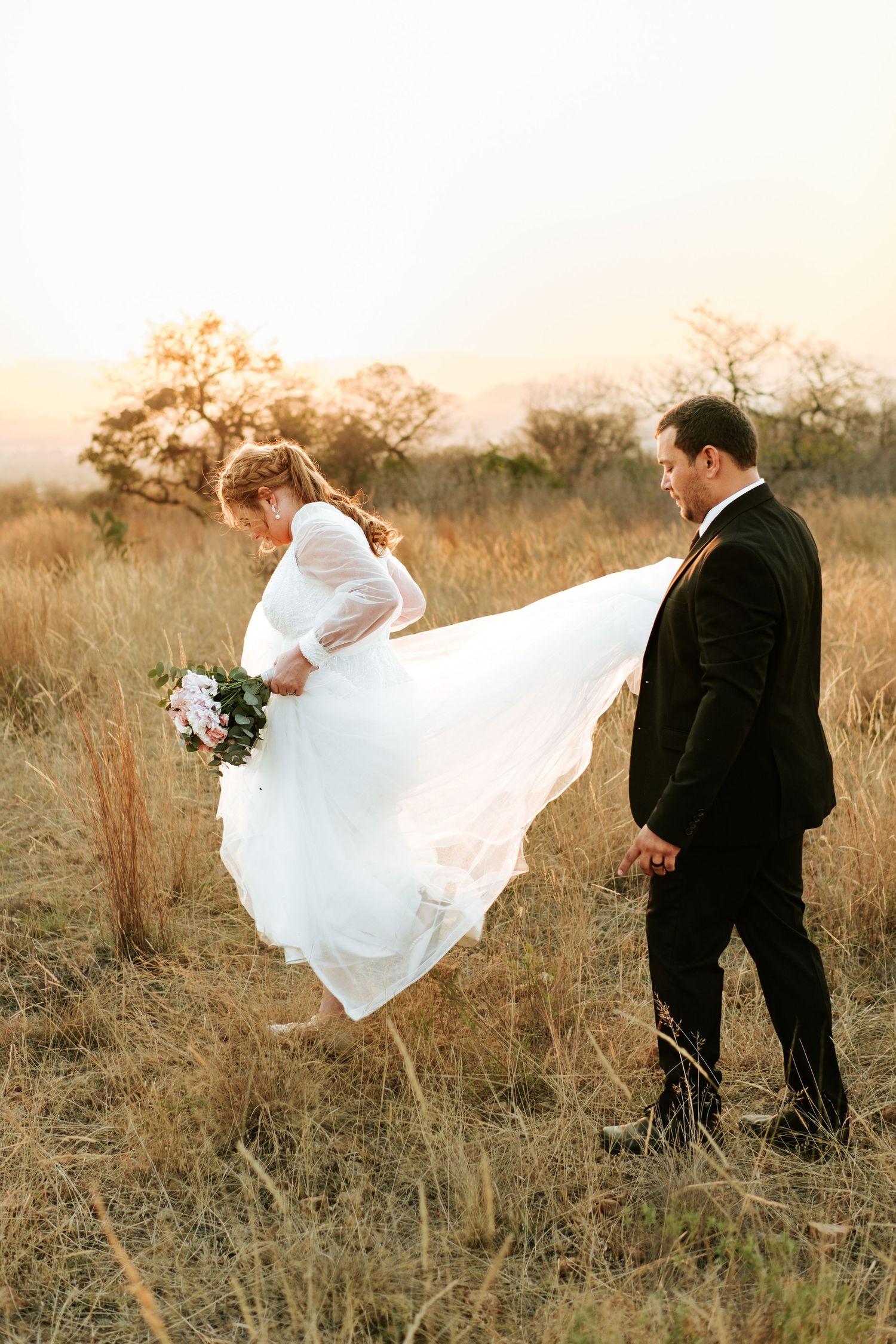 Bride lifts flowing white gown while groom watches during romantic sunset walk through dry grass field.