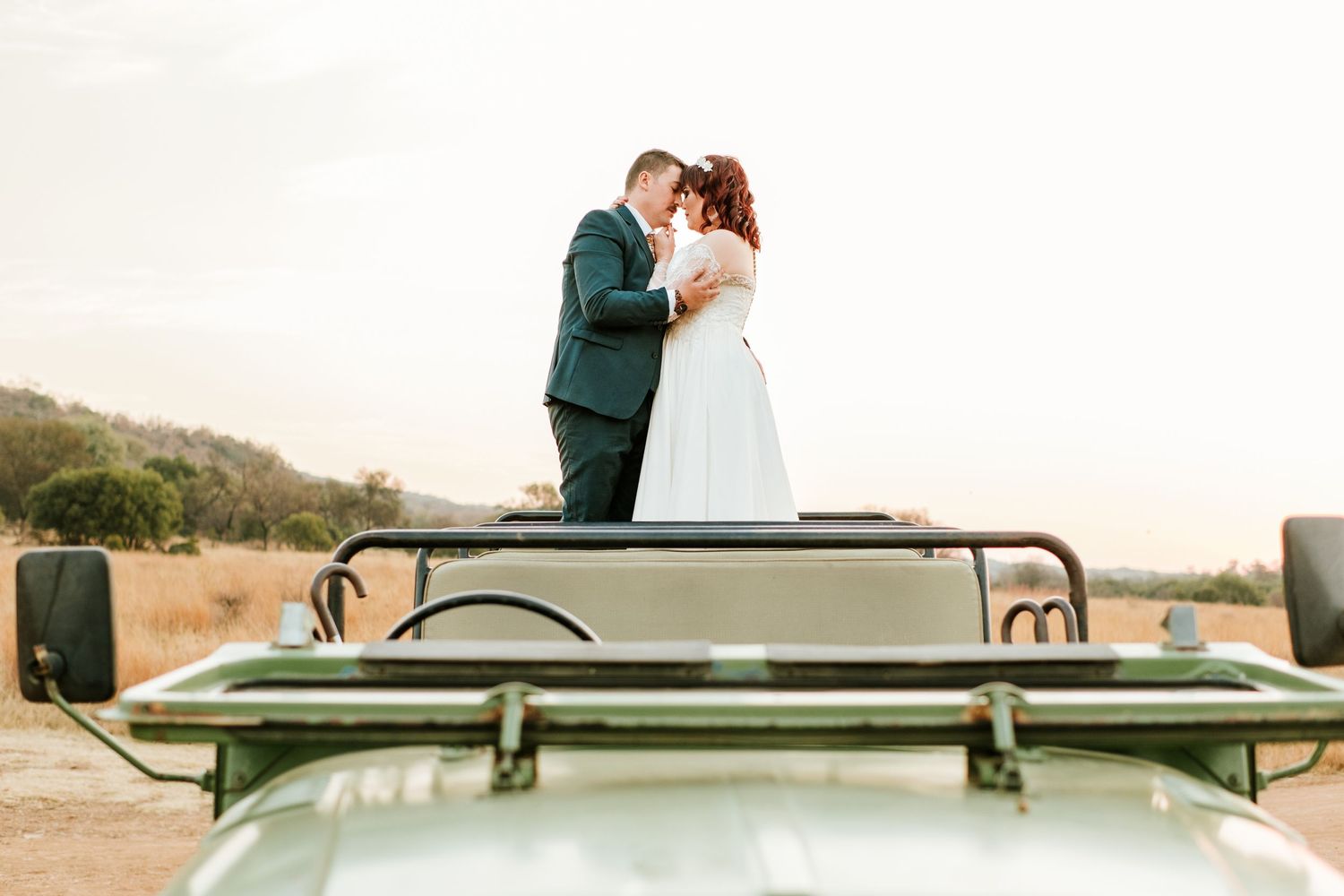 Bride and groom share tender kiss standing atop green safari jeep at golden hour sunset.