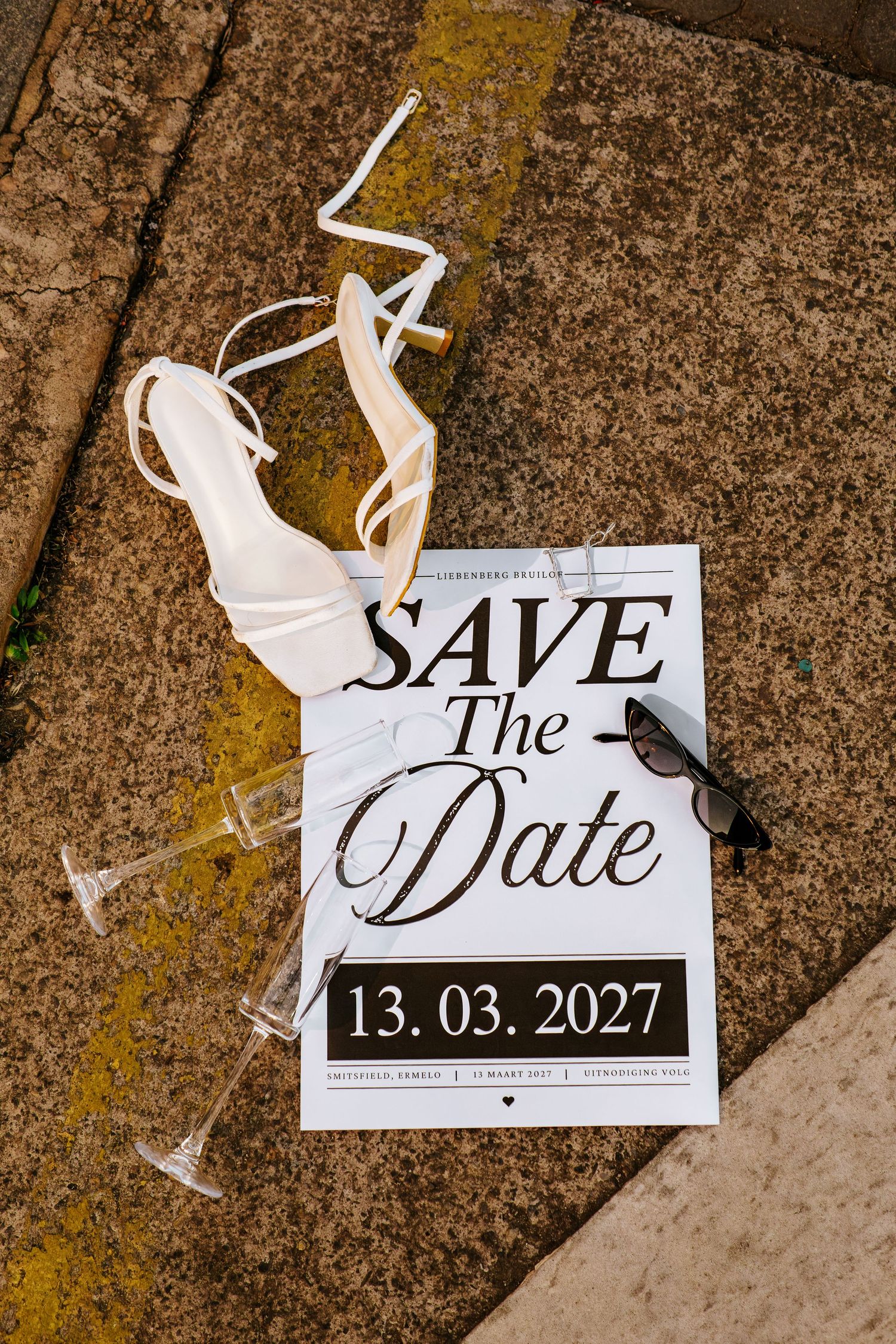 Wedding Save the Date card for 13.03.2027 with bridal heels, sunglasses and champagne flutes on stone surface.