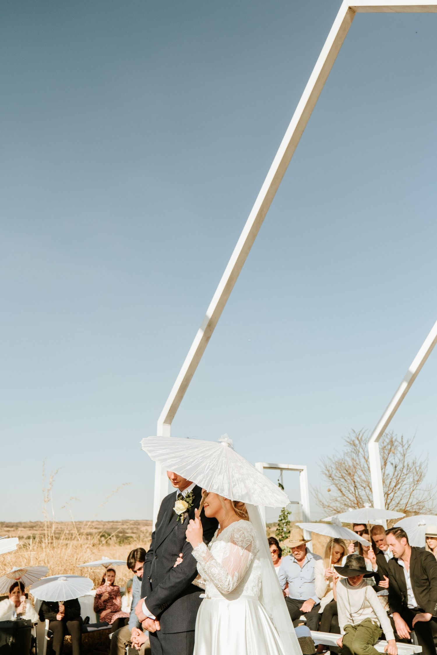 Bride and groom walk down outdoor aisle under white parasols during sunny wedding ceremony with seated guests.