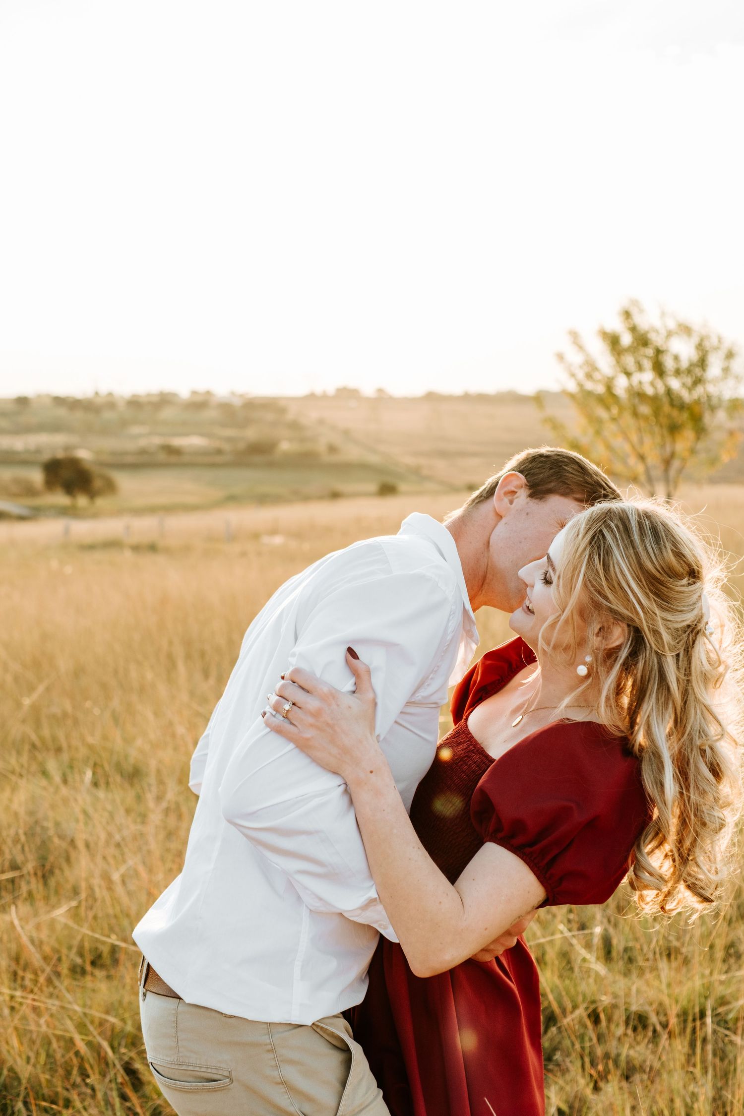 Couple sharing a romantic kiss in a golden field at sunset, woman in red dress embracing man in white shirt.
