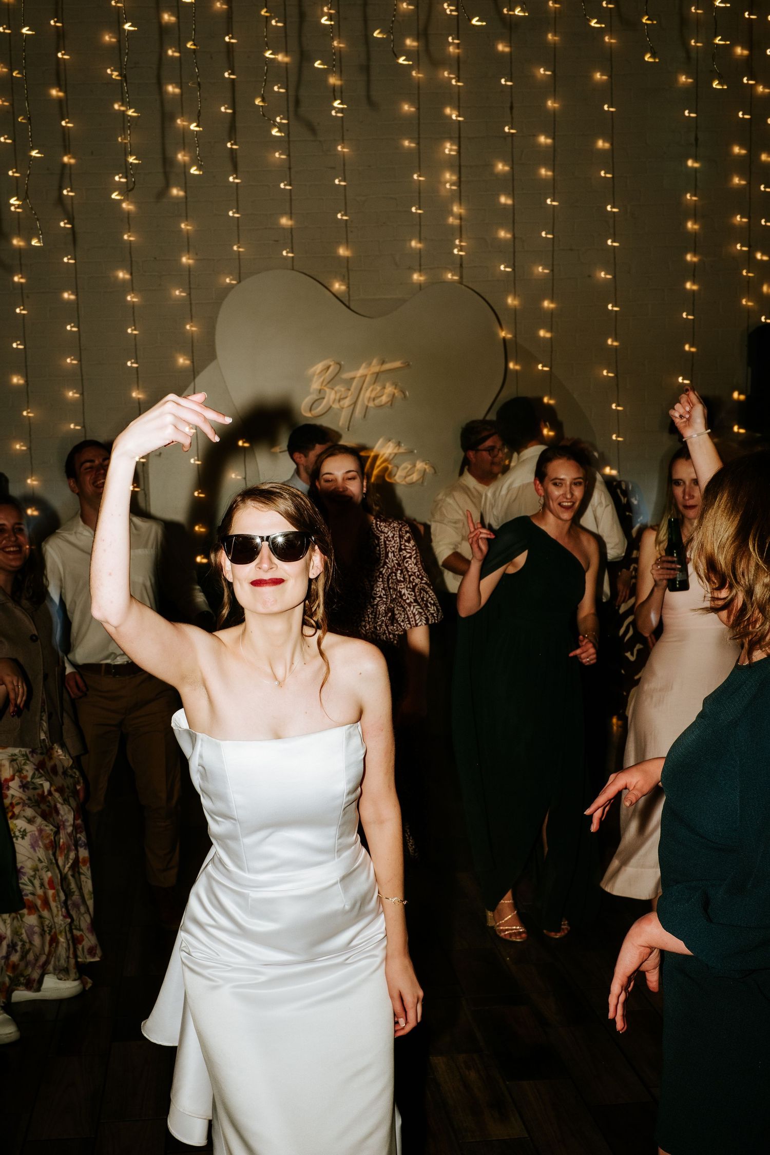 Bride in white strapless gown and sunglasses dancing joyfully at wedding reception with fairy light backdrop.