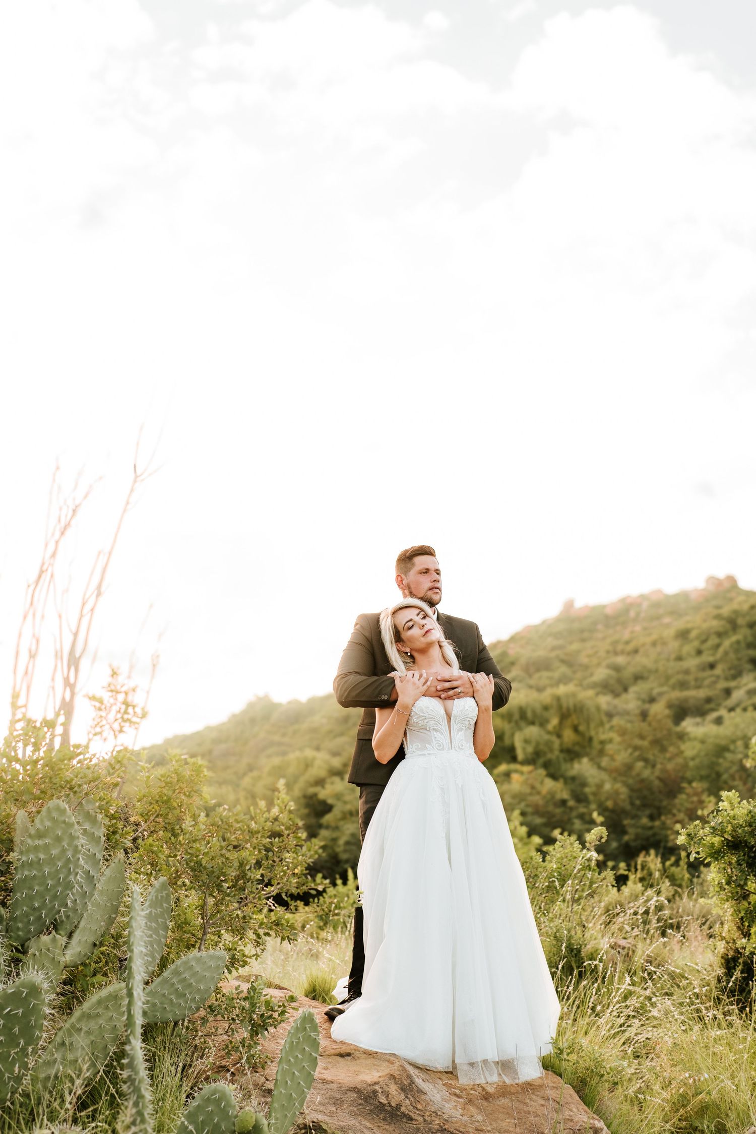 Groom embracing bride from behind on rocky hillside surrounded by lush green cacti and vegetation.