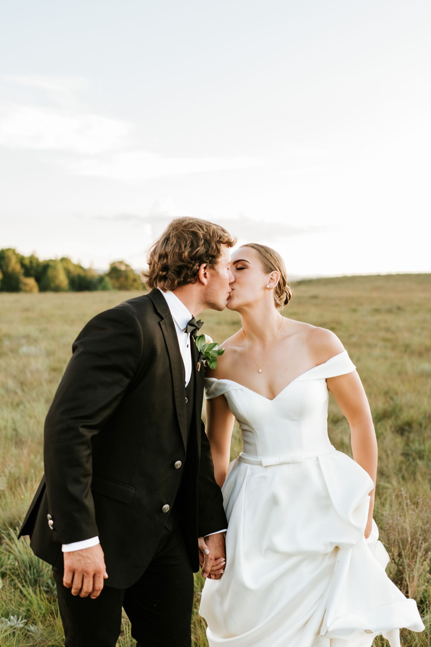 Bride and groom share a romantic kiss in an open meadow, bride in off-shoulder white gown.