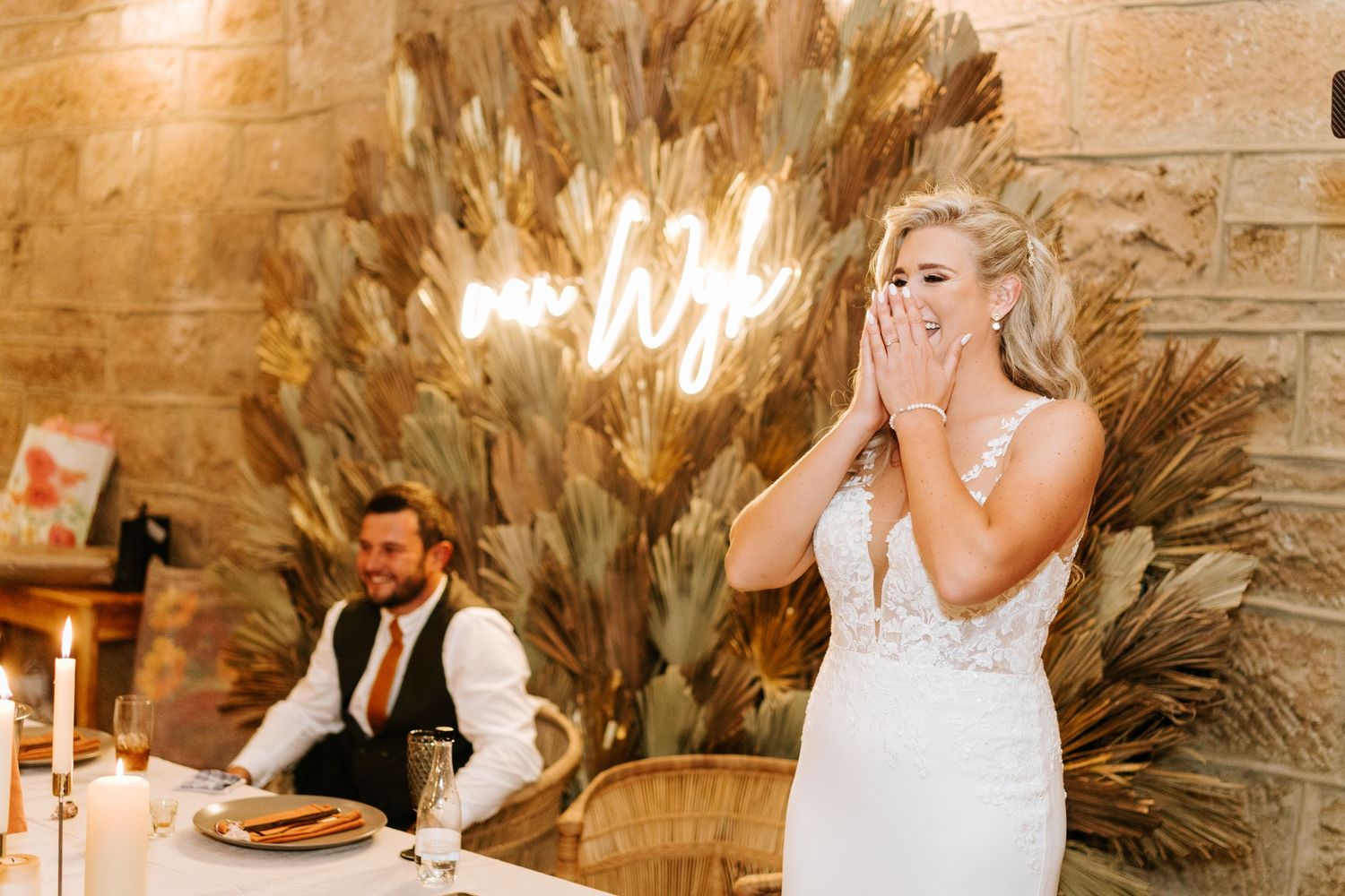 Emotional blonde bride in lace gown covers face with hands at rustic stone wall reception venue.