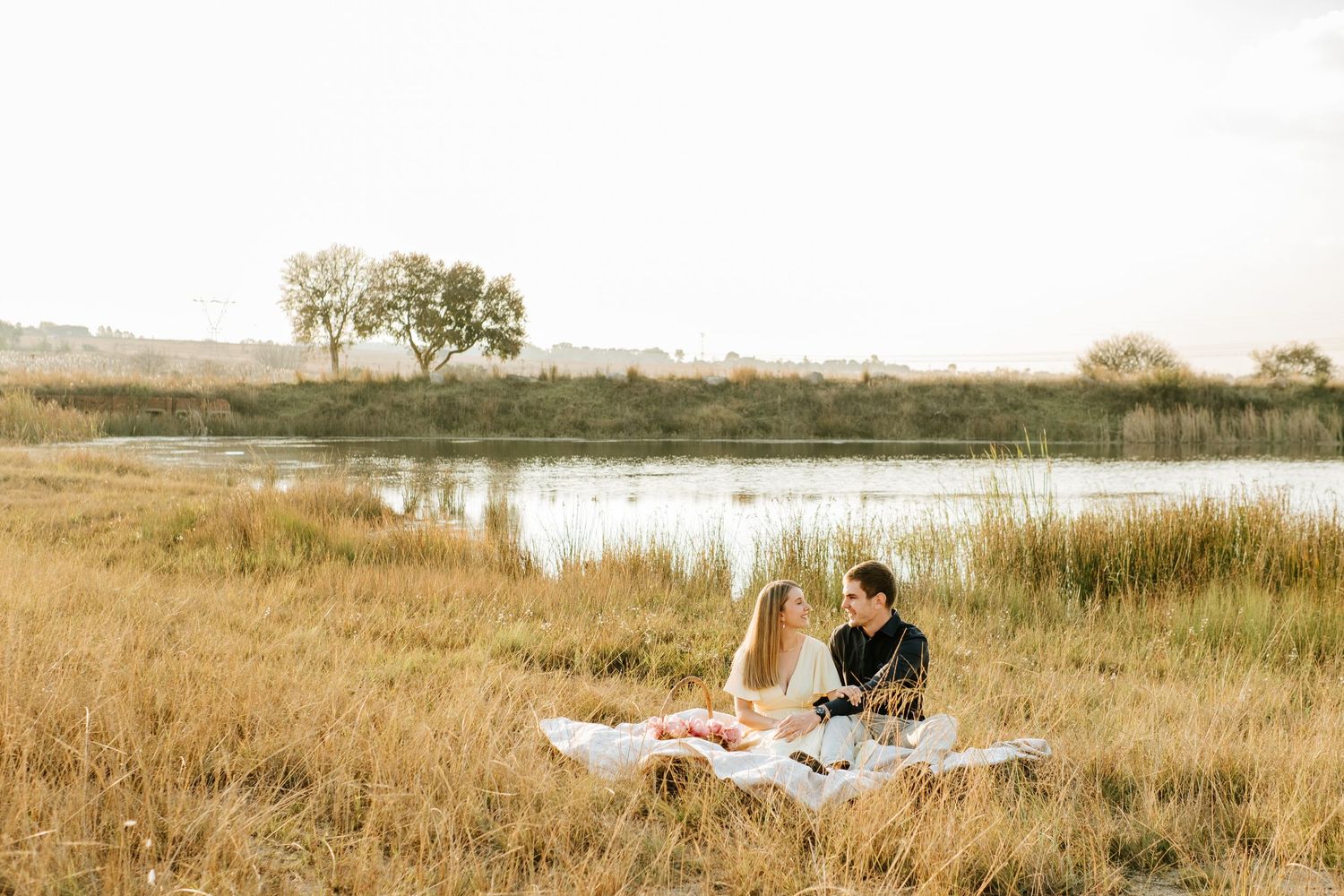 Romantic couple enjoying a picnic on a blanket in golden grass field beside a serene lake at sunset.