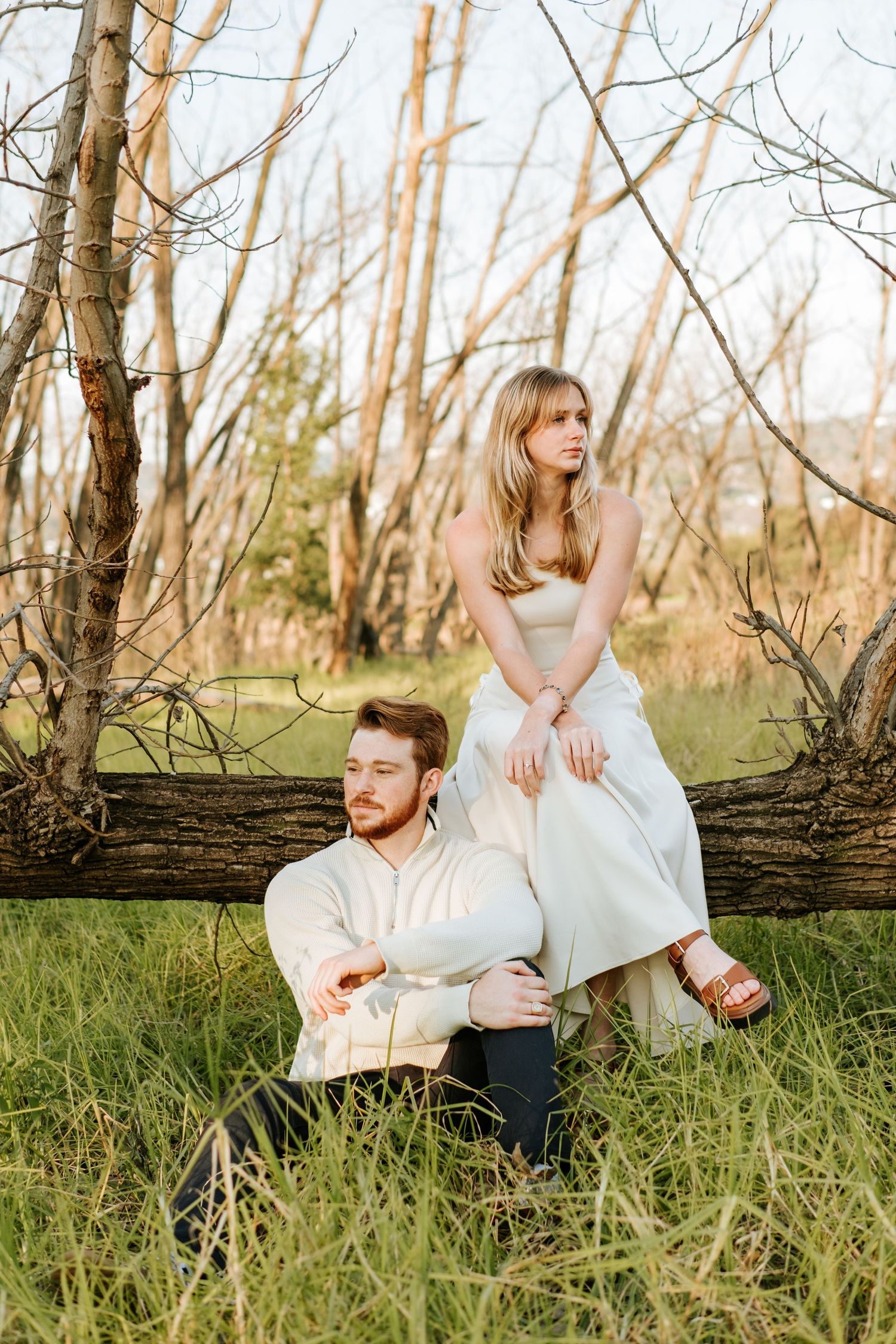 Couple posing by bare trees, woman in white dress sitting on log above man in white shirt.
