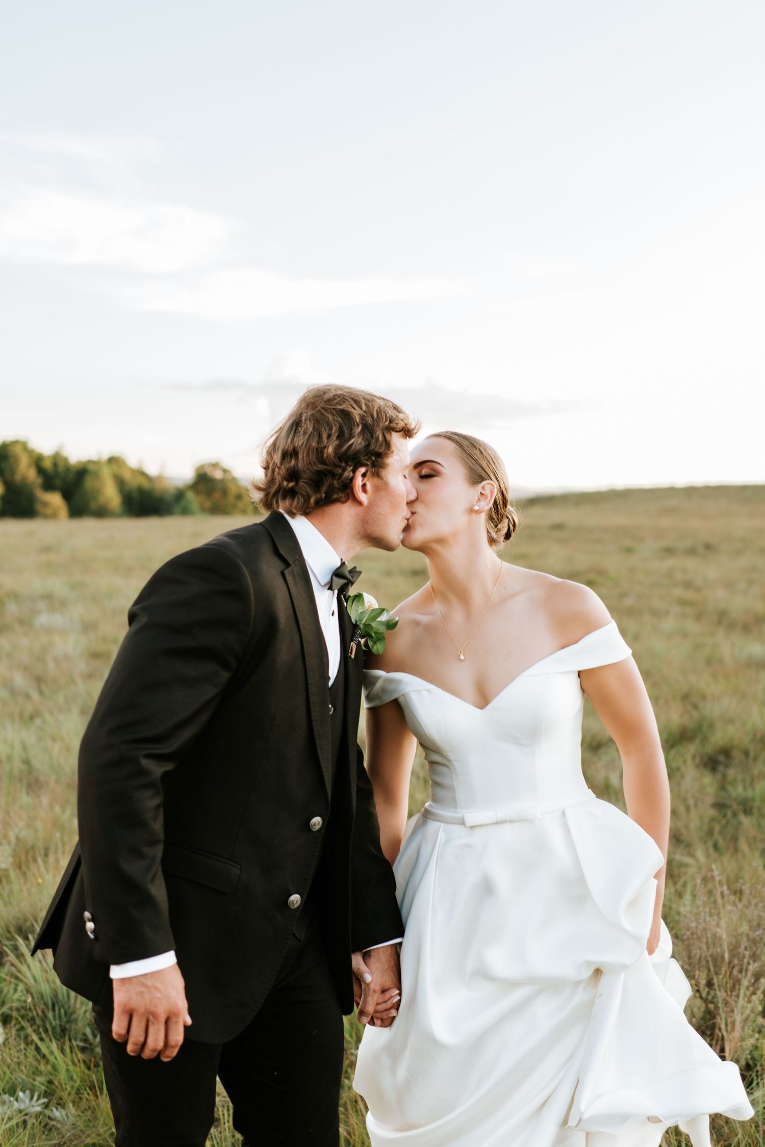 Bride and groom share a romantic kiss in an open field, bride wearing off-shoulder white wedding gown.