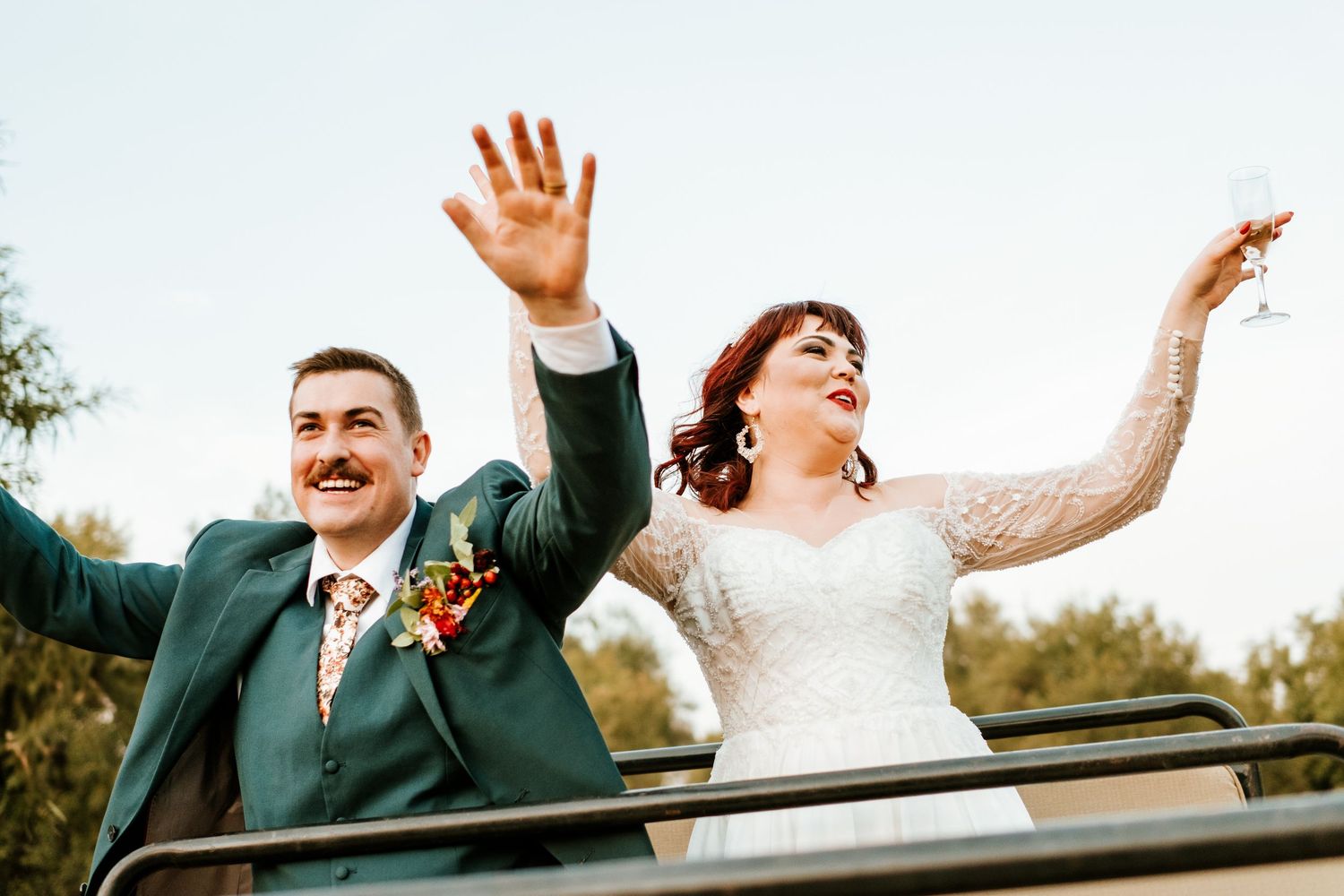 Joyful newlyweds waving from open-top vehicle, bride in lace gown and groom in teal suit with floral boutonniere.