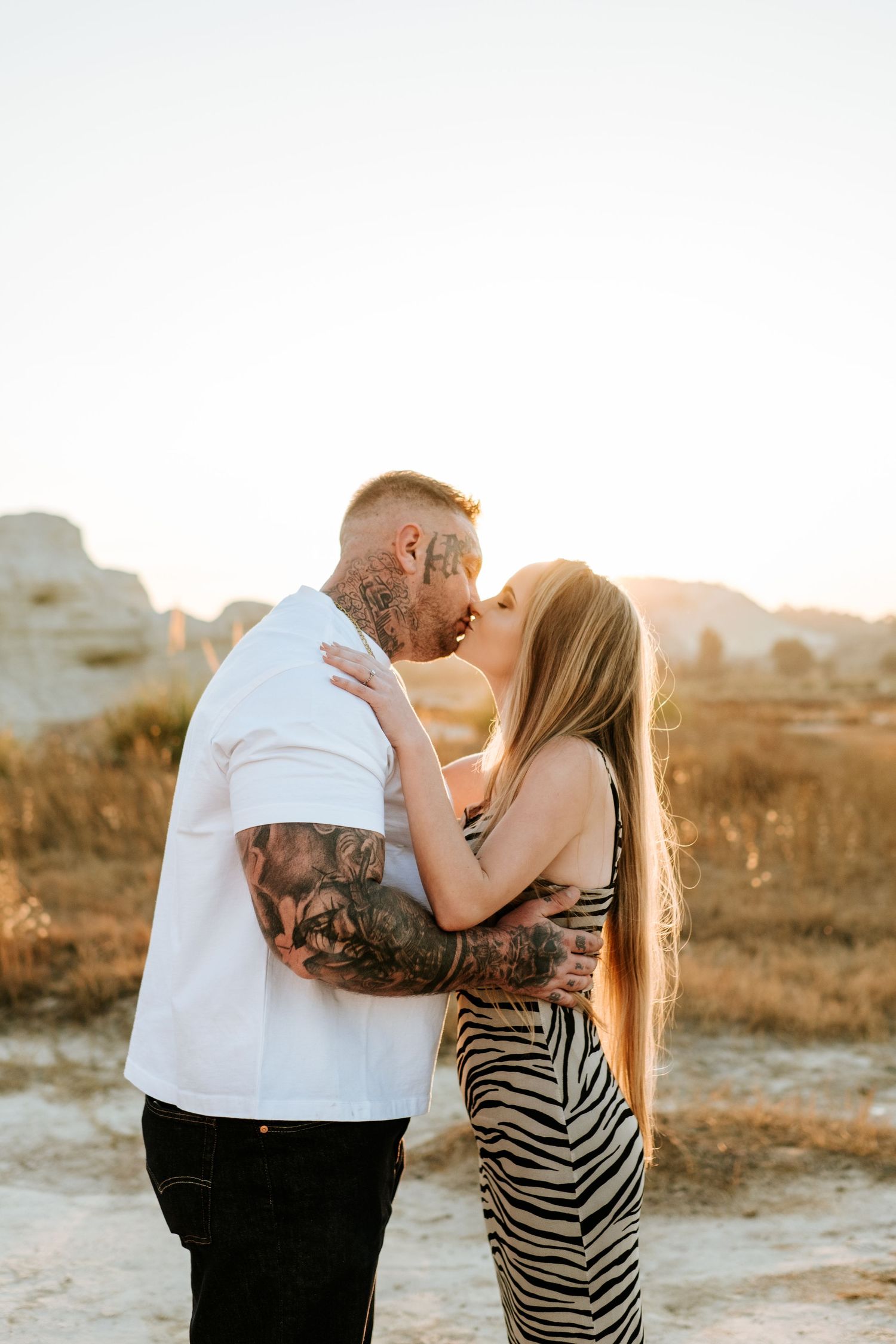 Tattooed groom and bride share a kiss in a sun-drenched field with rocky landscape behind them.