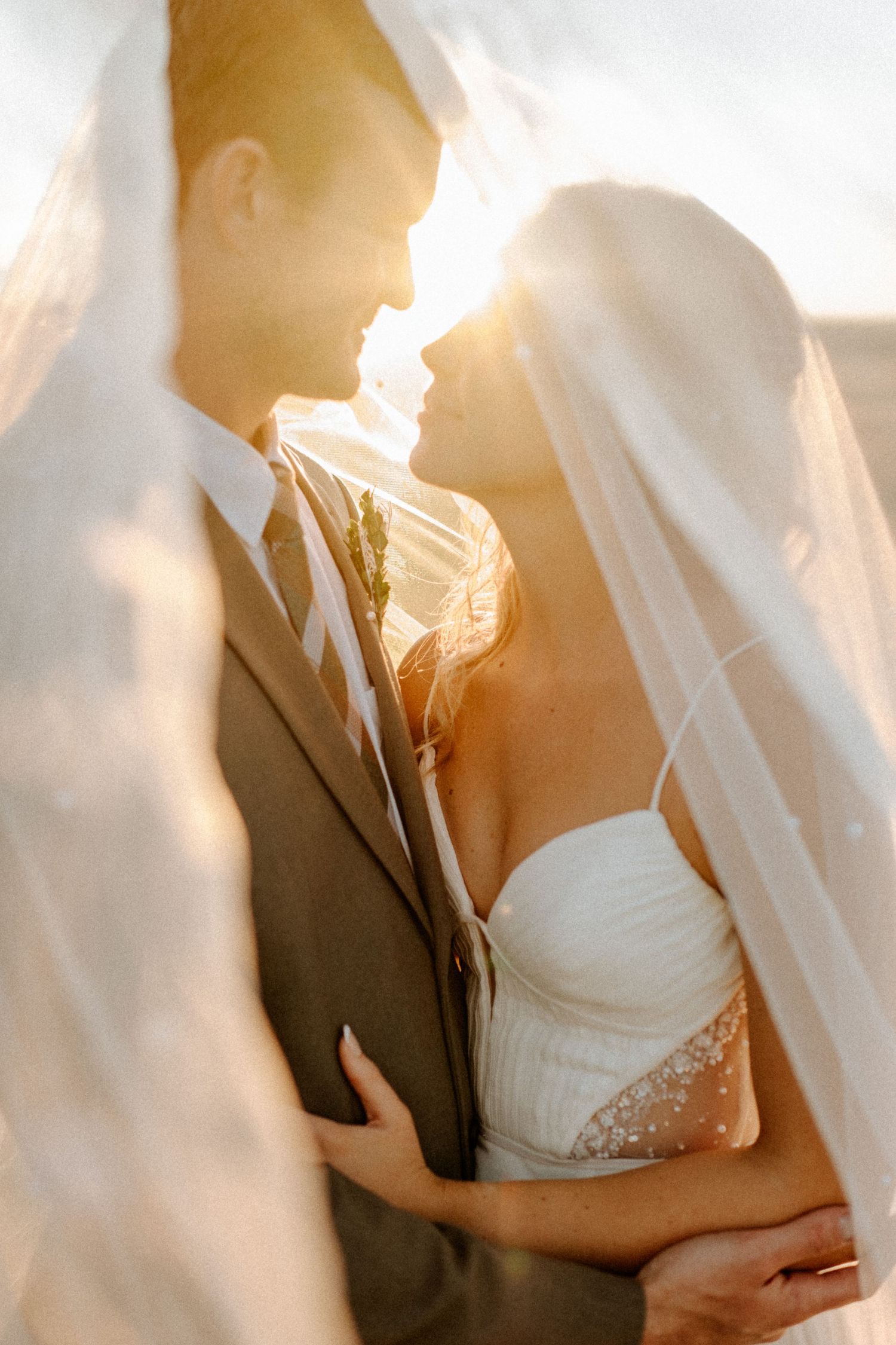 Bride and groom share an intimate moment under a flowing veil with golden sunset backlight.