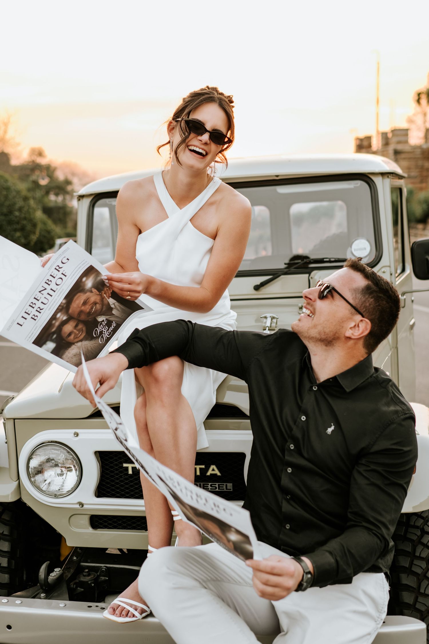 Happy engaged couple laughing together with Save the Date cards beside a vintage white Toyota FJ40 at sunset.