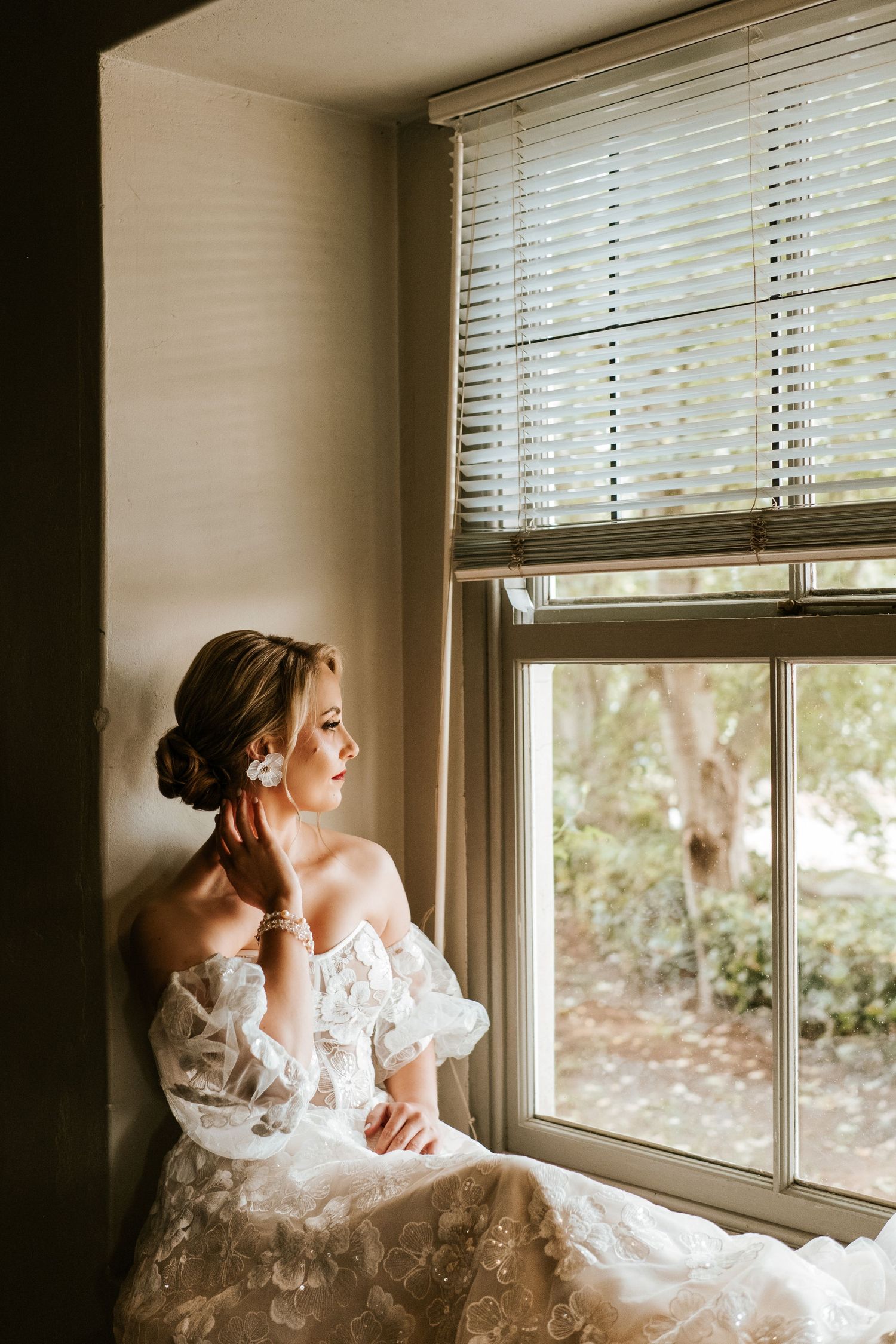 Elegant bride in off-shoulder lace gown sits pensively by a sunlit window in soft warm light.
