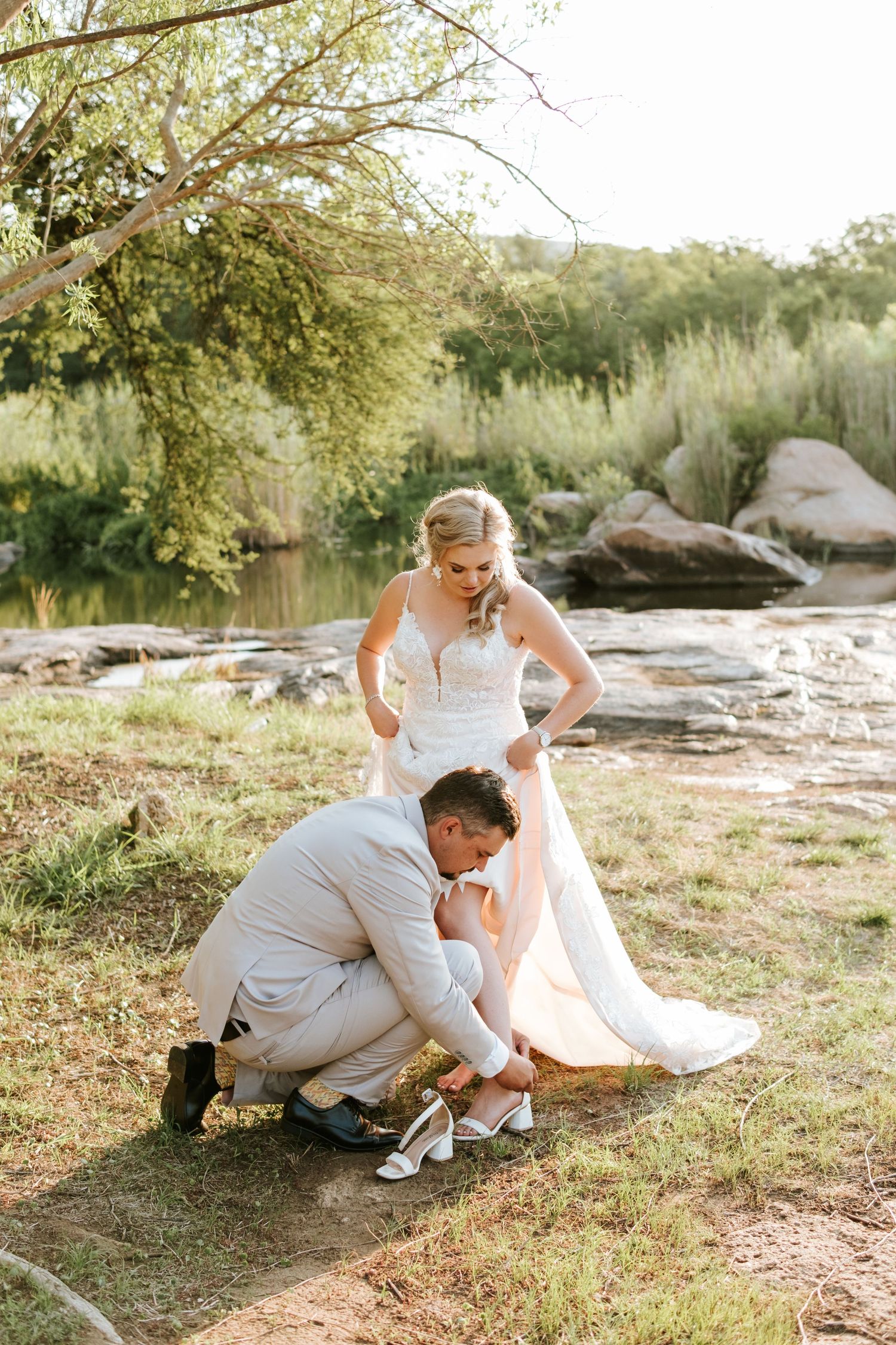 Groom kneeling to adjust bride's wedding shoe by a scenic riverside in golden hour light.