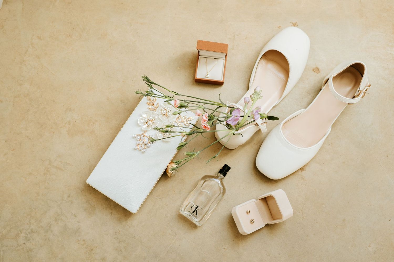 Elegant bridal accessories flatlay featuring white heels, perfume, ring boxes, and delicate flowers.