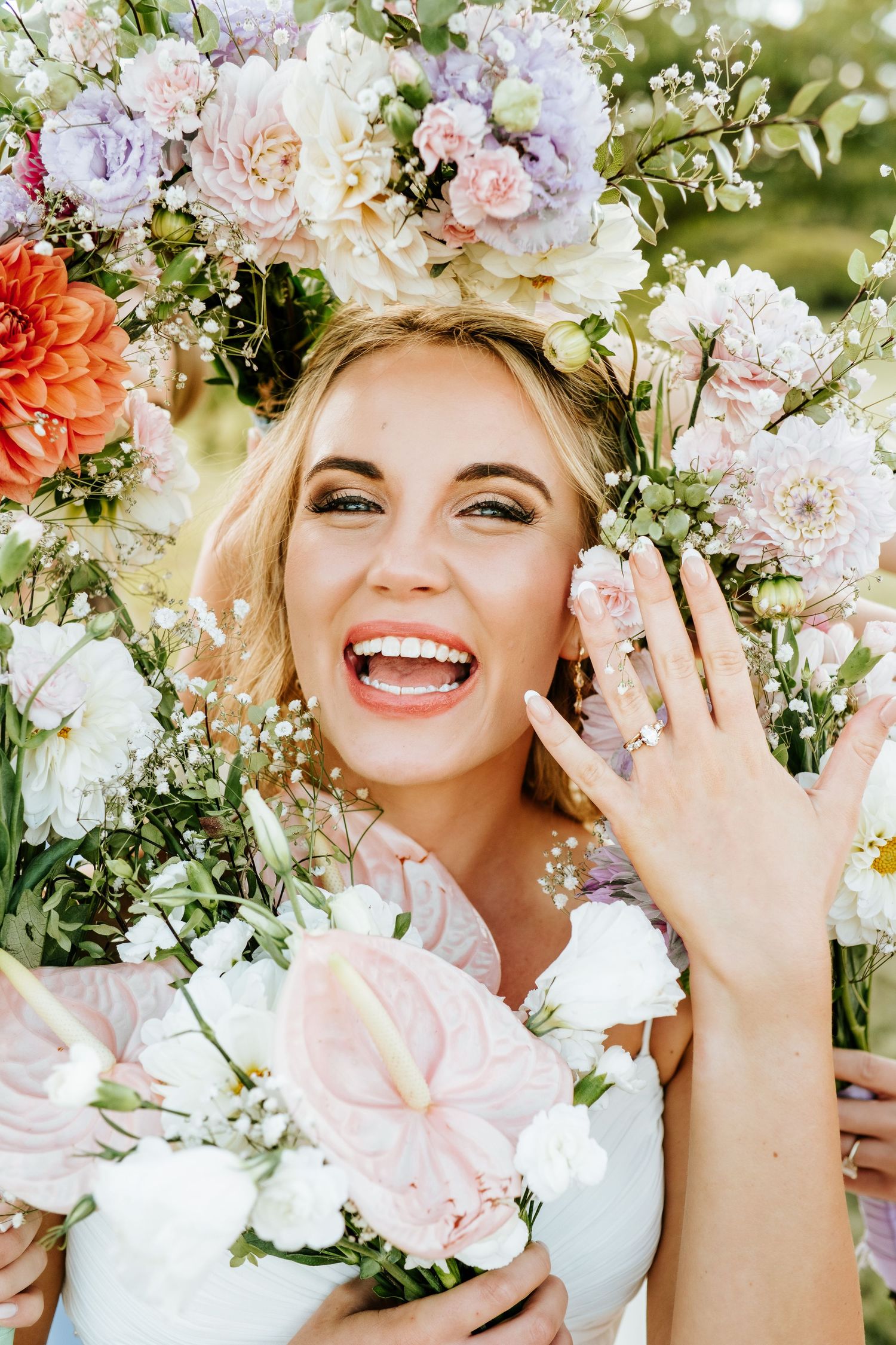 Radiant smiling bride surrounded by colorful floral arch displaying her engagement ring at outdoor wedding.