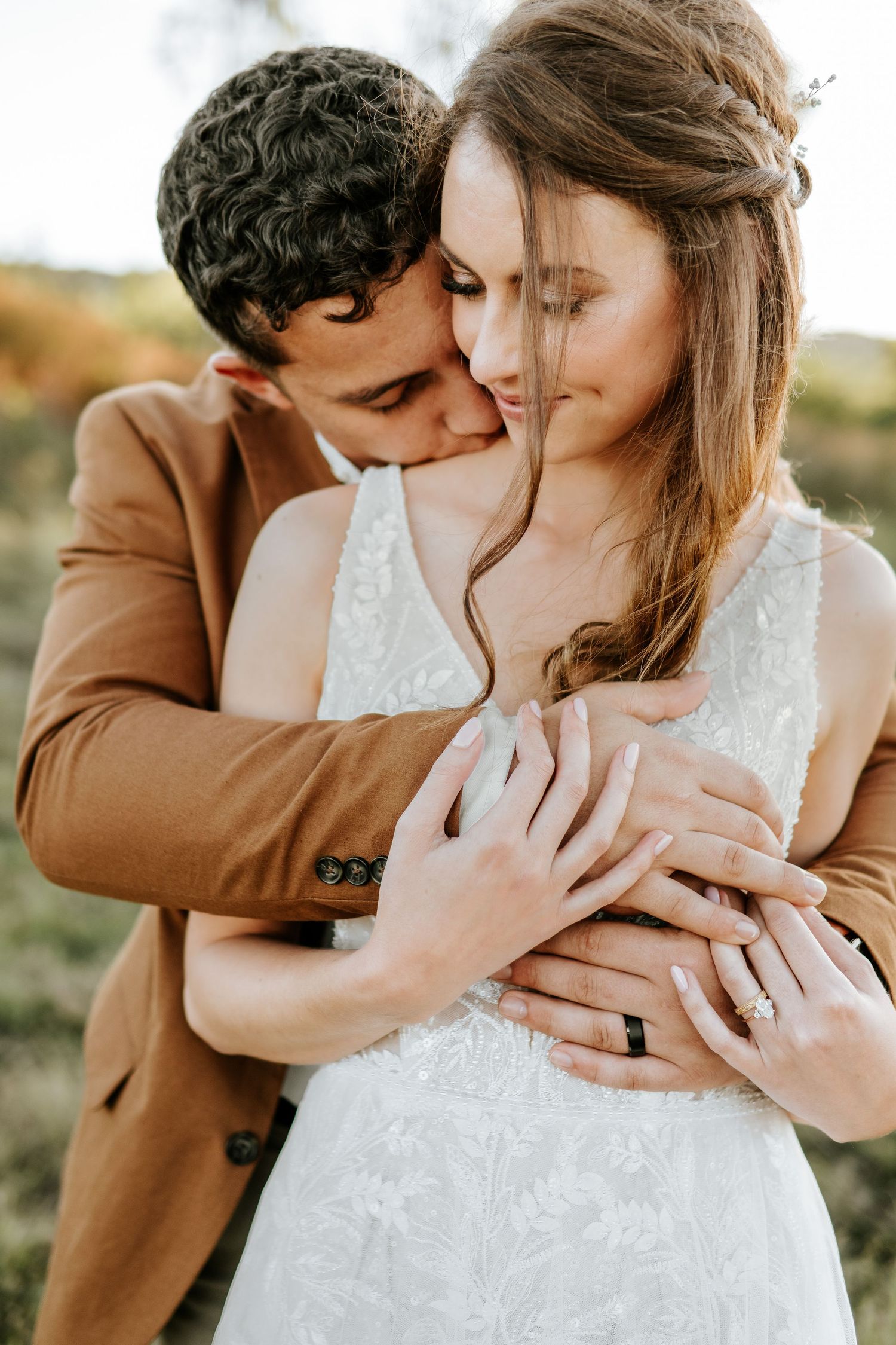 Groom in tan suit embracing bride from behind in outdoor setting, both wearing wedding rings.