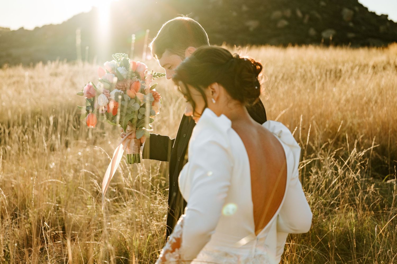 Bride and groom share a romantic moment in golden field, bride wearing elegant open-back wedding gown holding colorful bouquet.