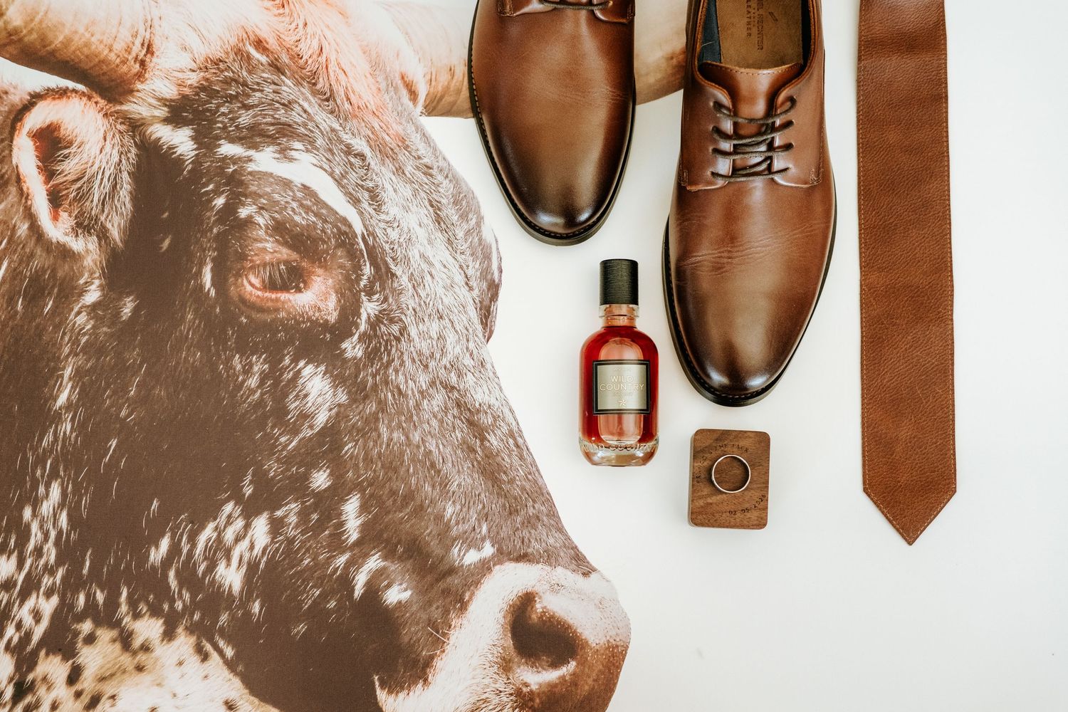 Brown leather wedding shoes, tie, cologne, and ring styled with a longhorn cattle print.