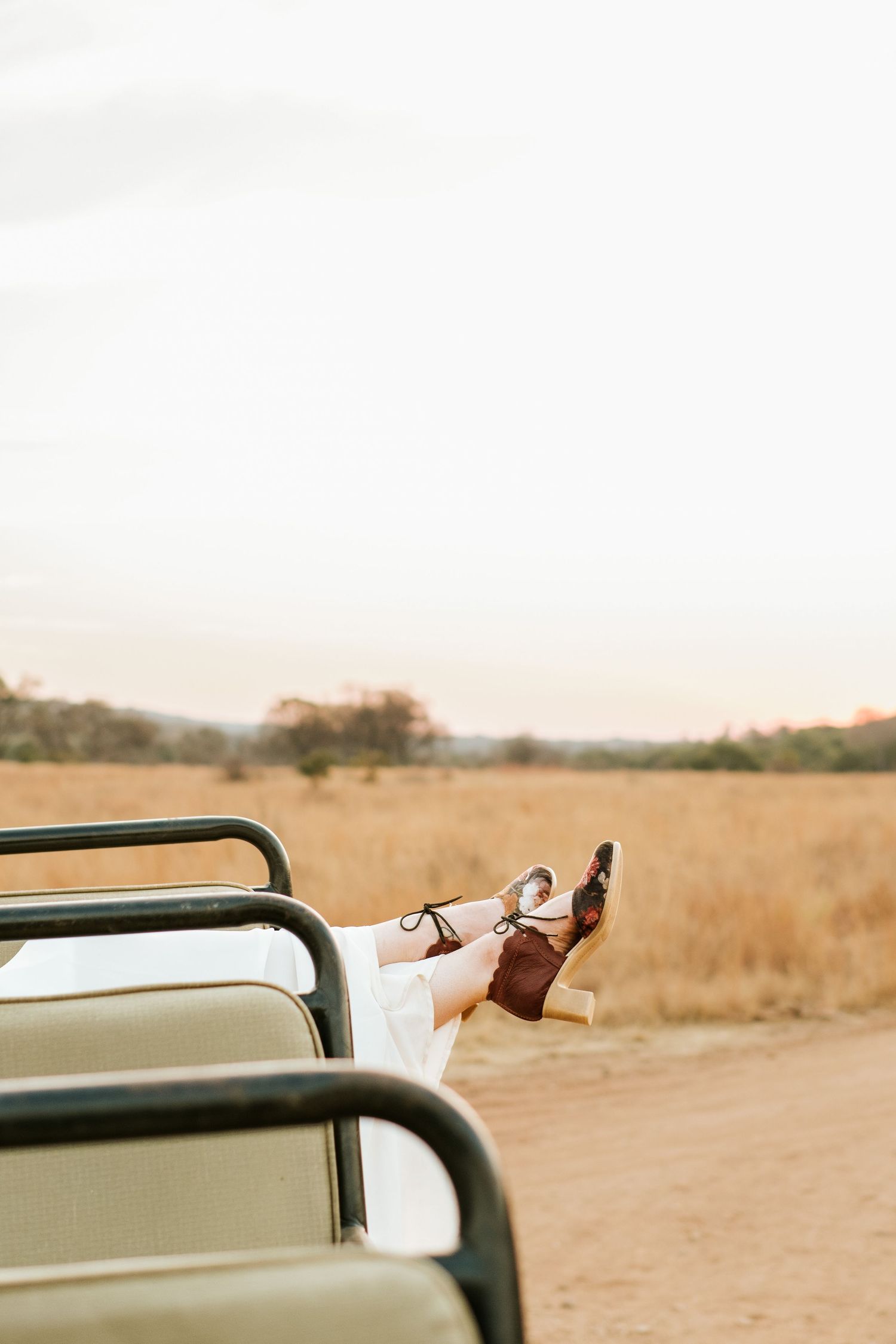 Bride resting legs over safari vehicle seat wearing brown heeled sandals in golden savanna landscape.