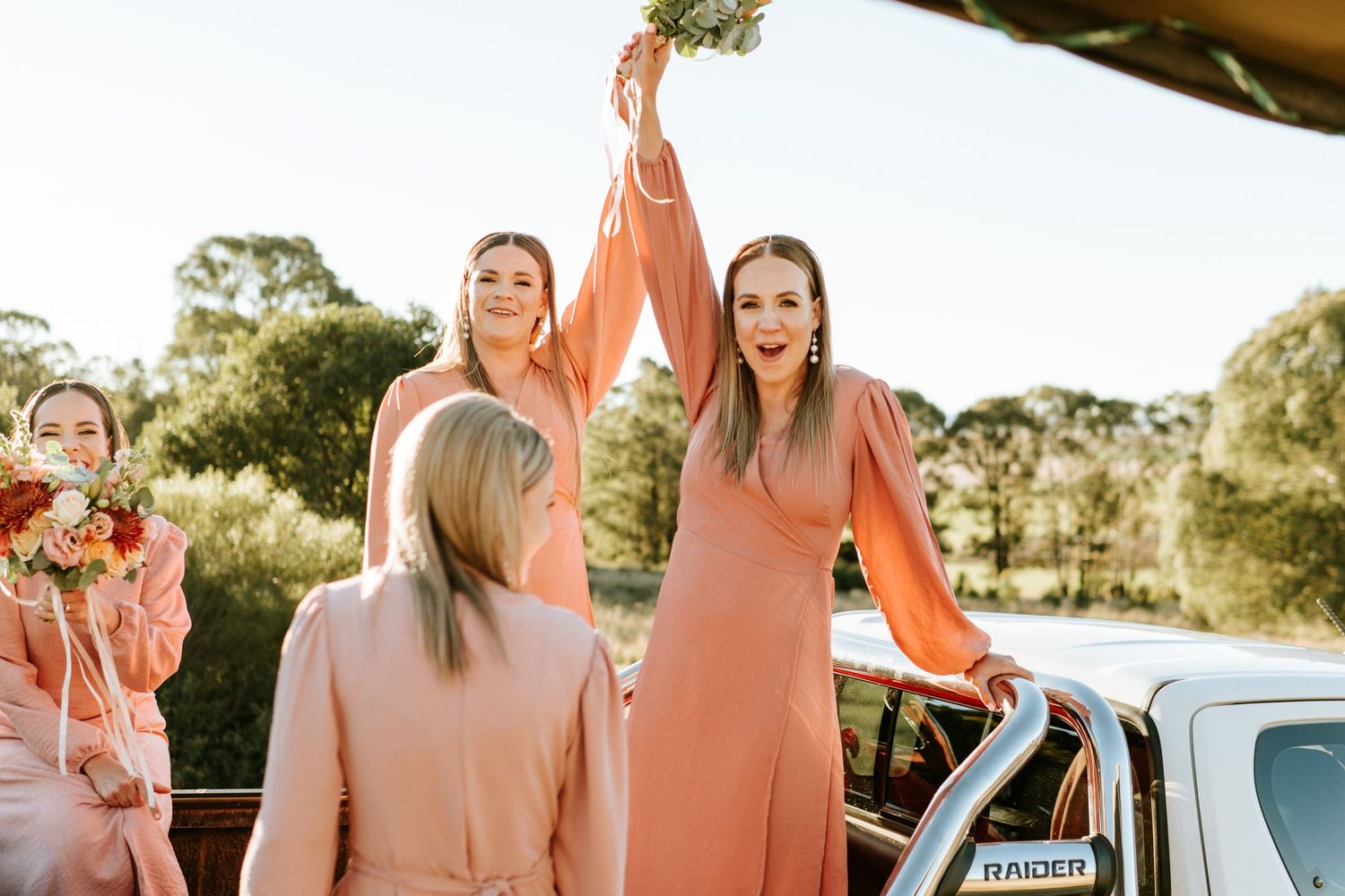 Bridesmaids in blush pink dresses celebrate joyfully beside a truck in sunny outdoors.