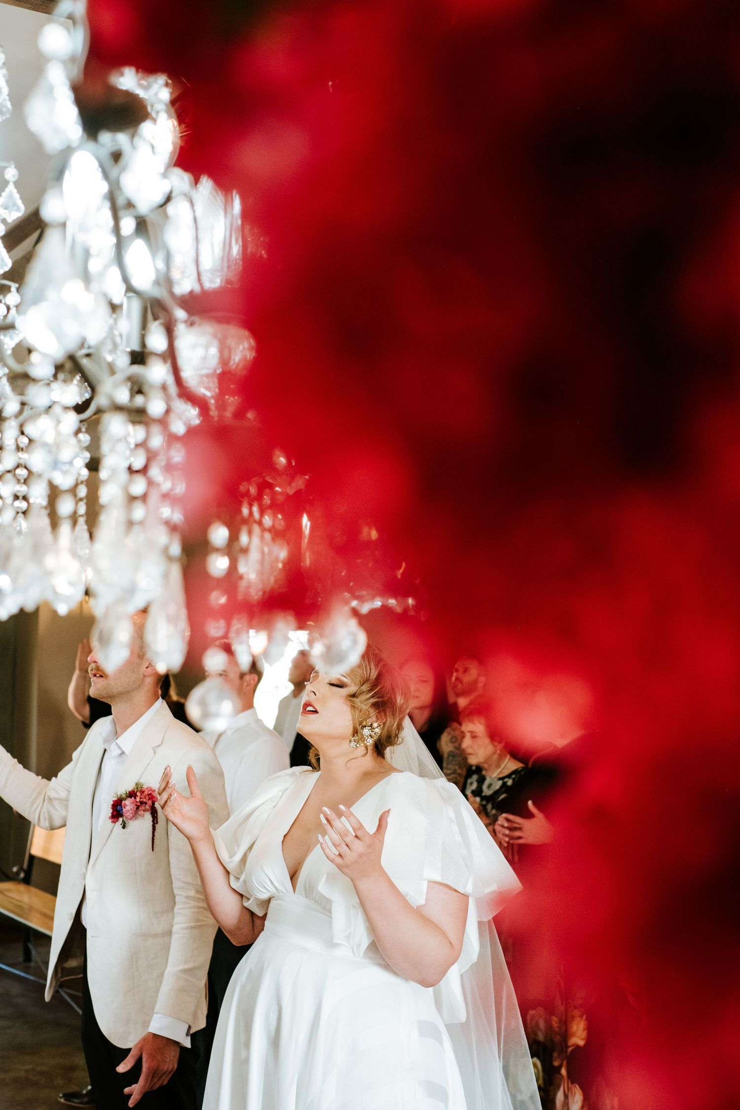 Bride and groom dancing joyfully beneath crystal chandeliers framed by vibrant red floral decor.