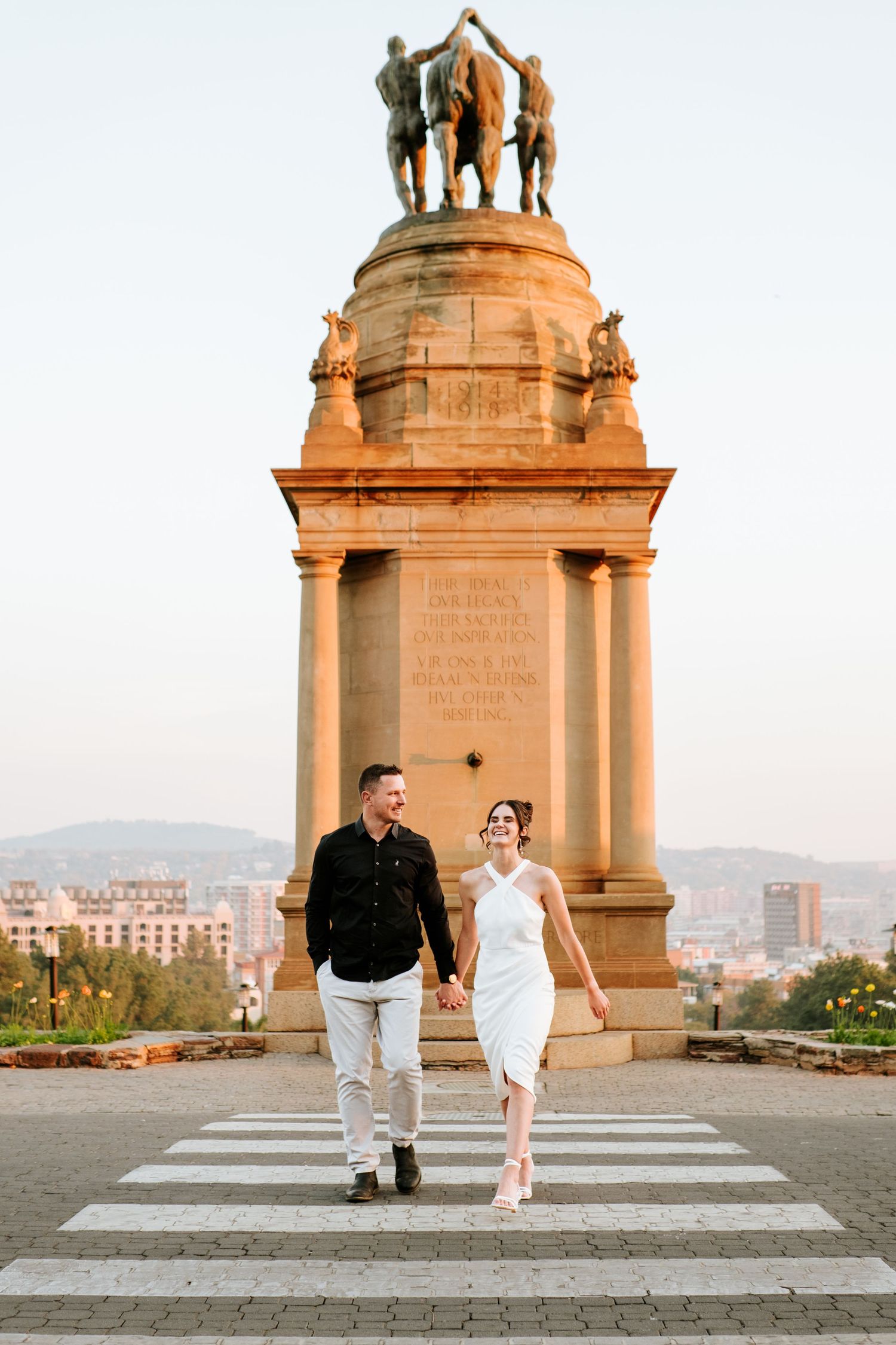 Smiling couple walks hand in hand across a zebra crossing in front of a grand monument at dusk.