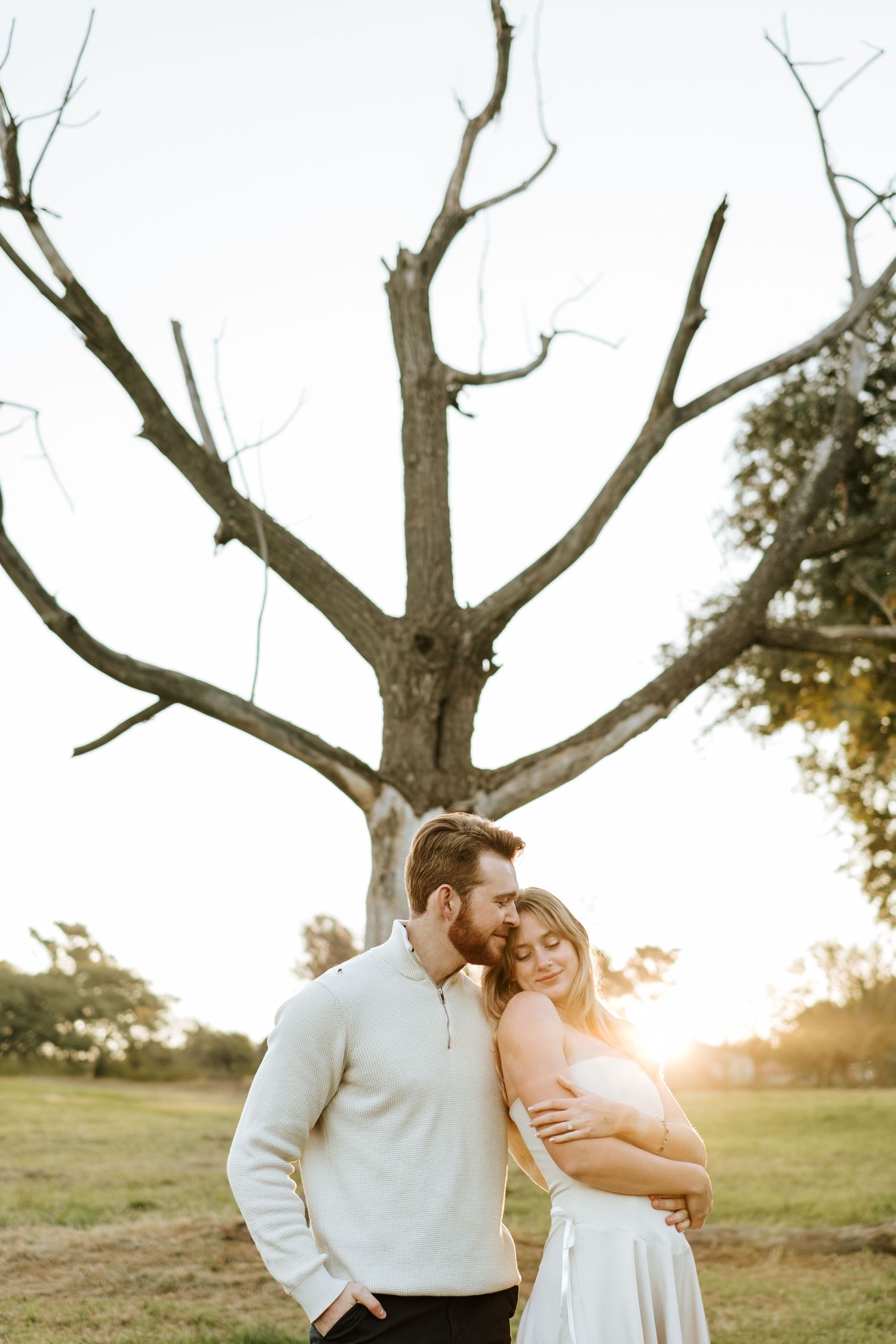 Couple embracing tenderly before a bare tree at golden sunset, man in white sweater with blonde woman.