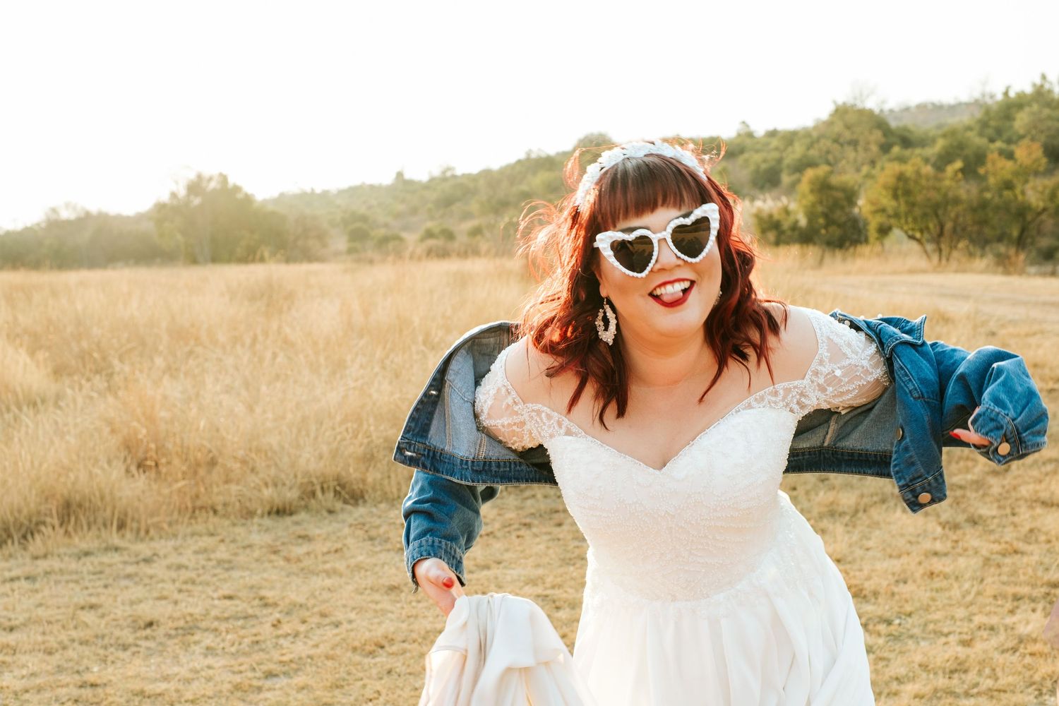 Joyful bride in white lace gown wearing heart sunglasses and denim jacket in a golden field.