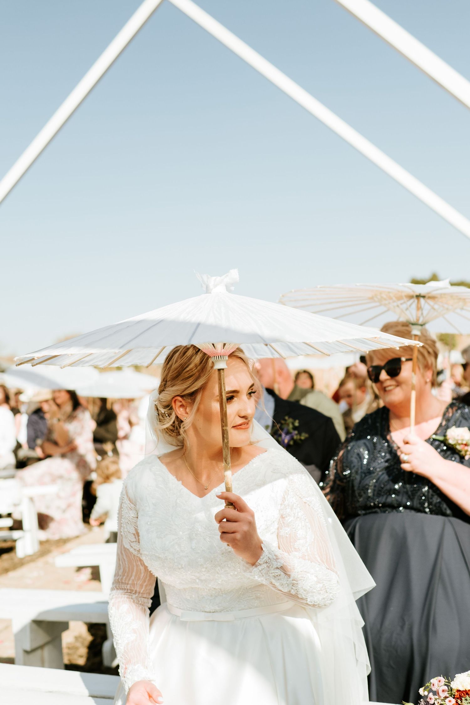 Blonde bride holding a white parasol umbrella while walking down the aisle at an outdoor ceremony.