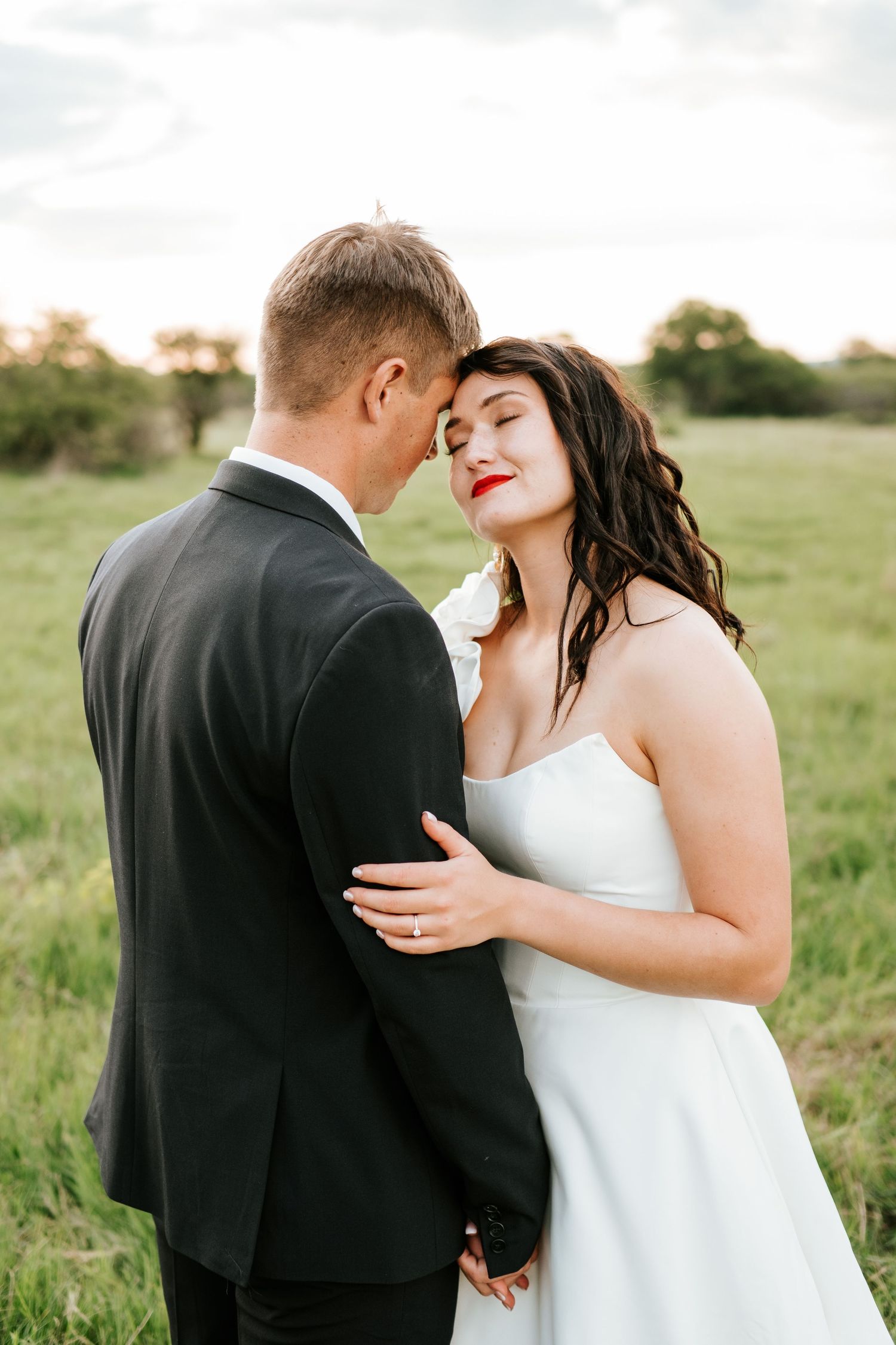 Bride with red lips embraces groom tenderly in a green field during soft golden hour light.