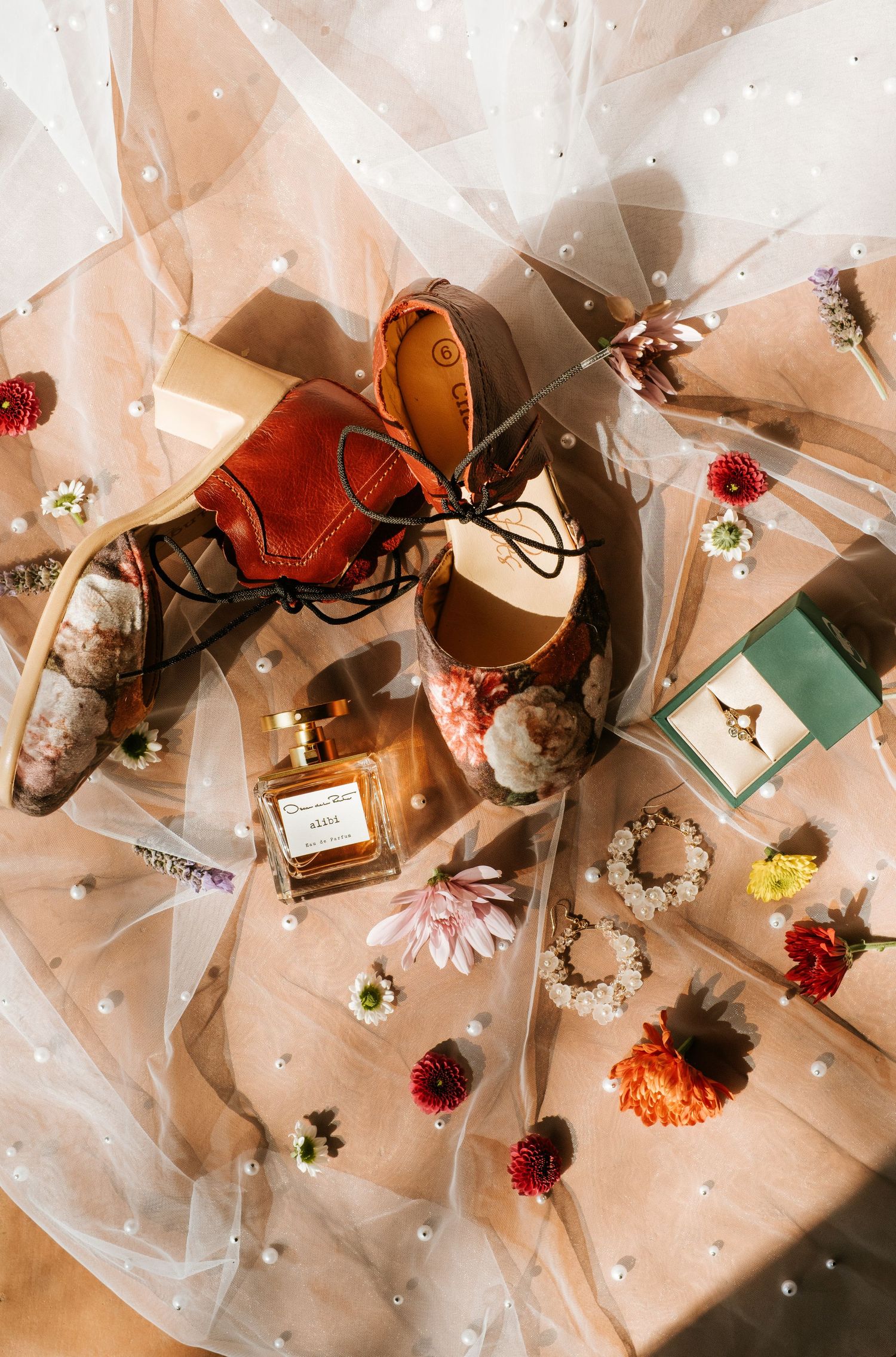 Flat lay of bridal accessories including floral heels, perfume, ring box, and flowers on pearl-dotted tulle.